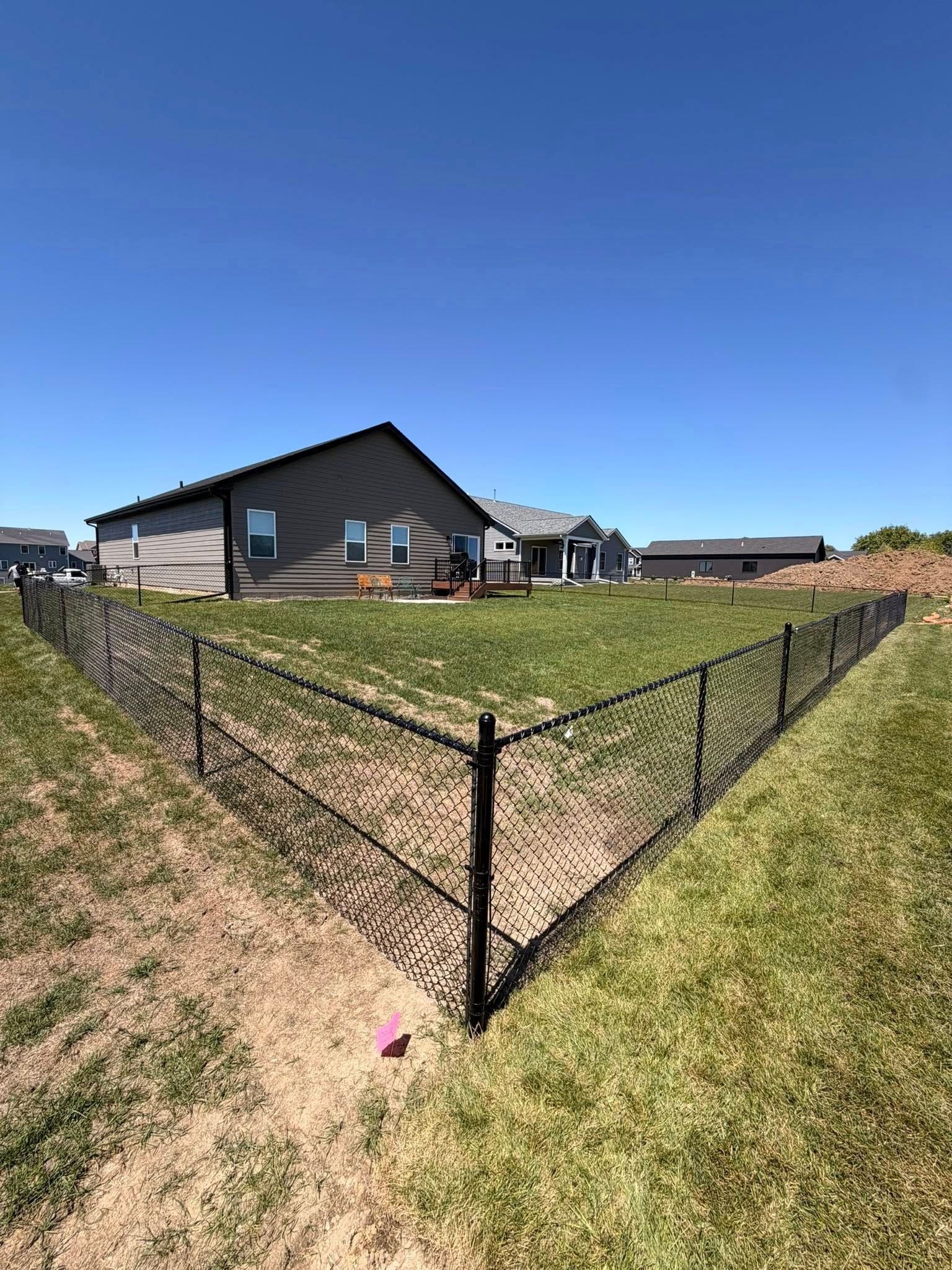 Suburban yard with black chain-link fence around a grassy lot beside a house under a clear blue sky