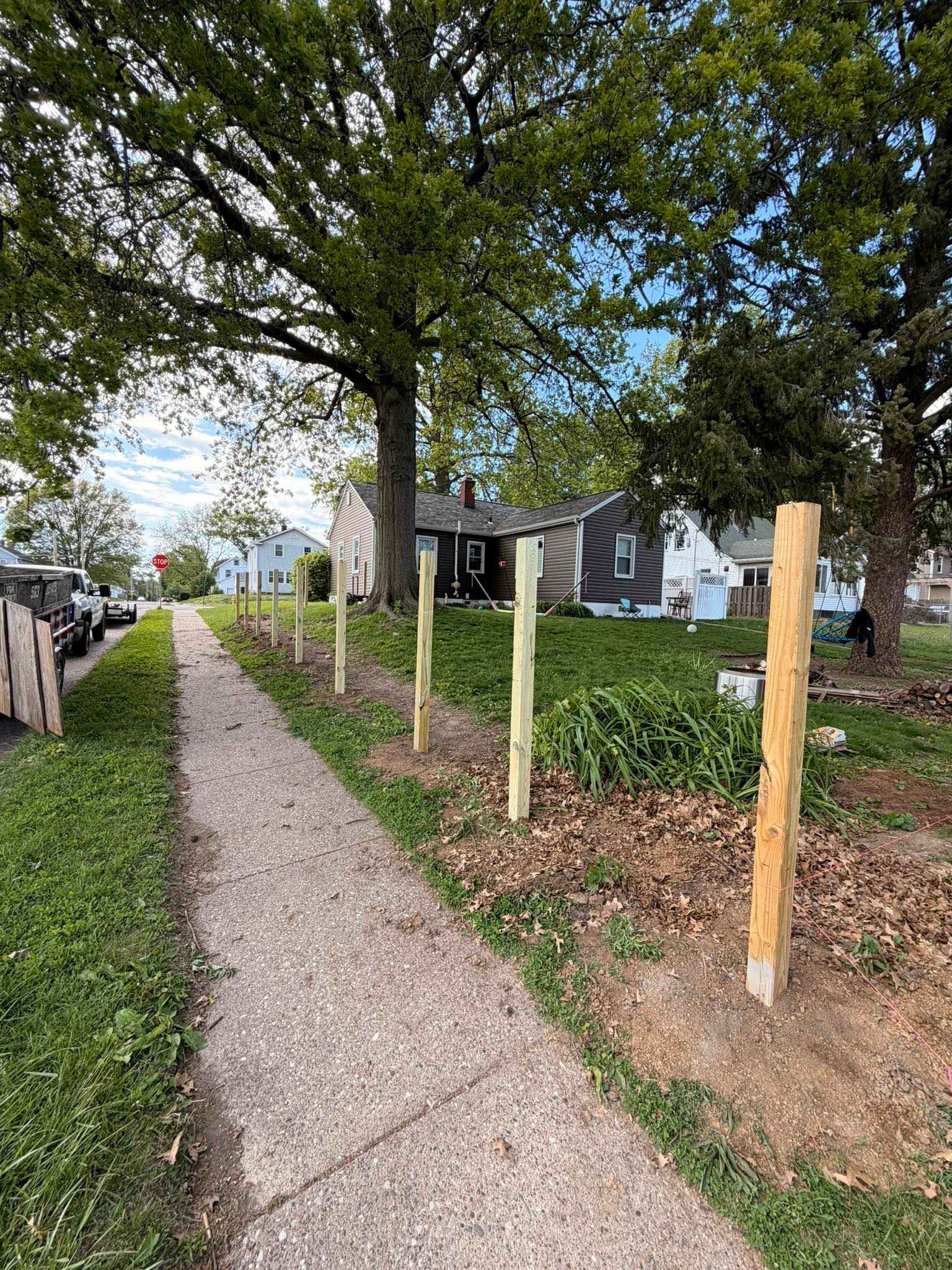 Sidewalk under trees beside a house, with wooden fence posts and a small yard.