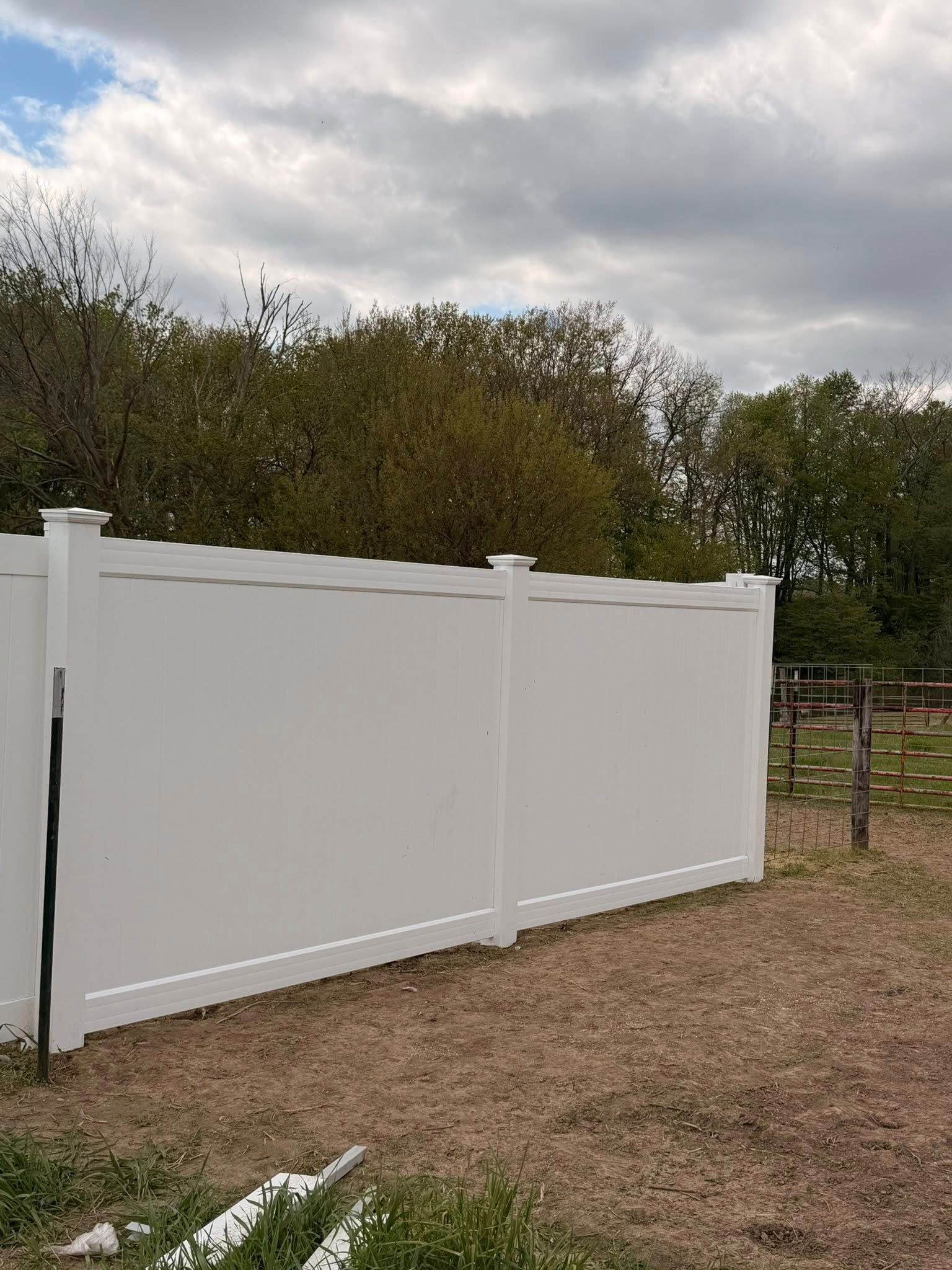 White vinyl fence beside a dirt path under cloudy sky, with trees and a gate in the background