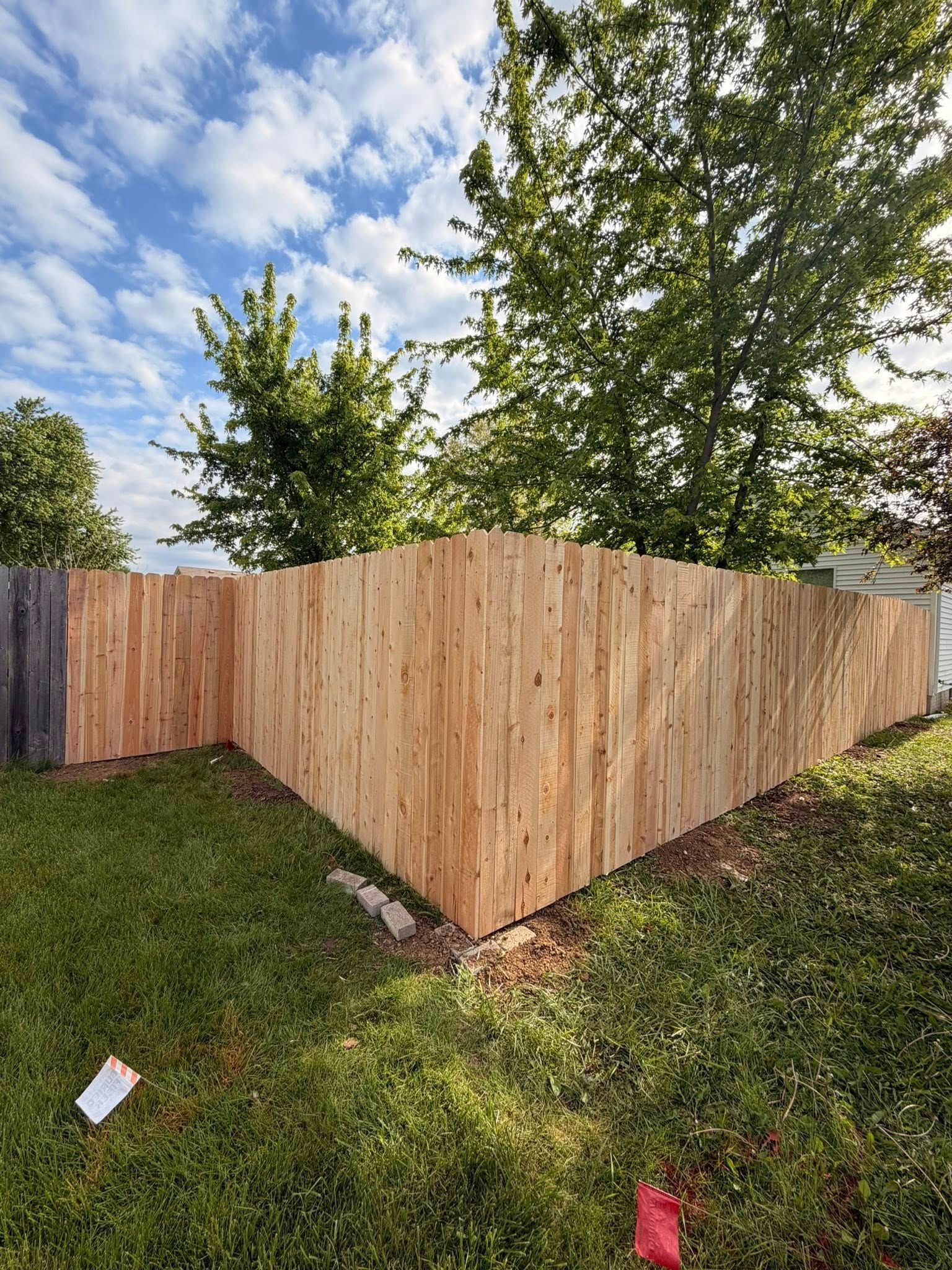 Wooden corner fence in a grassy yard beneath leafy trees and a partly cloudy sky.