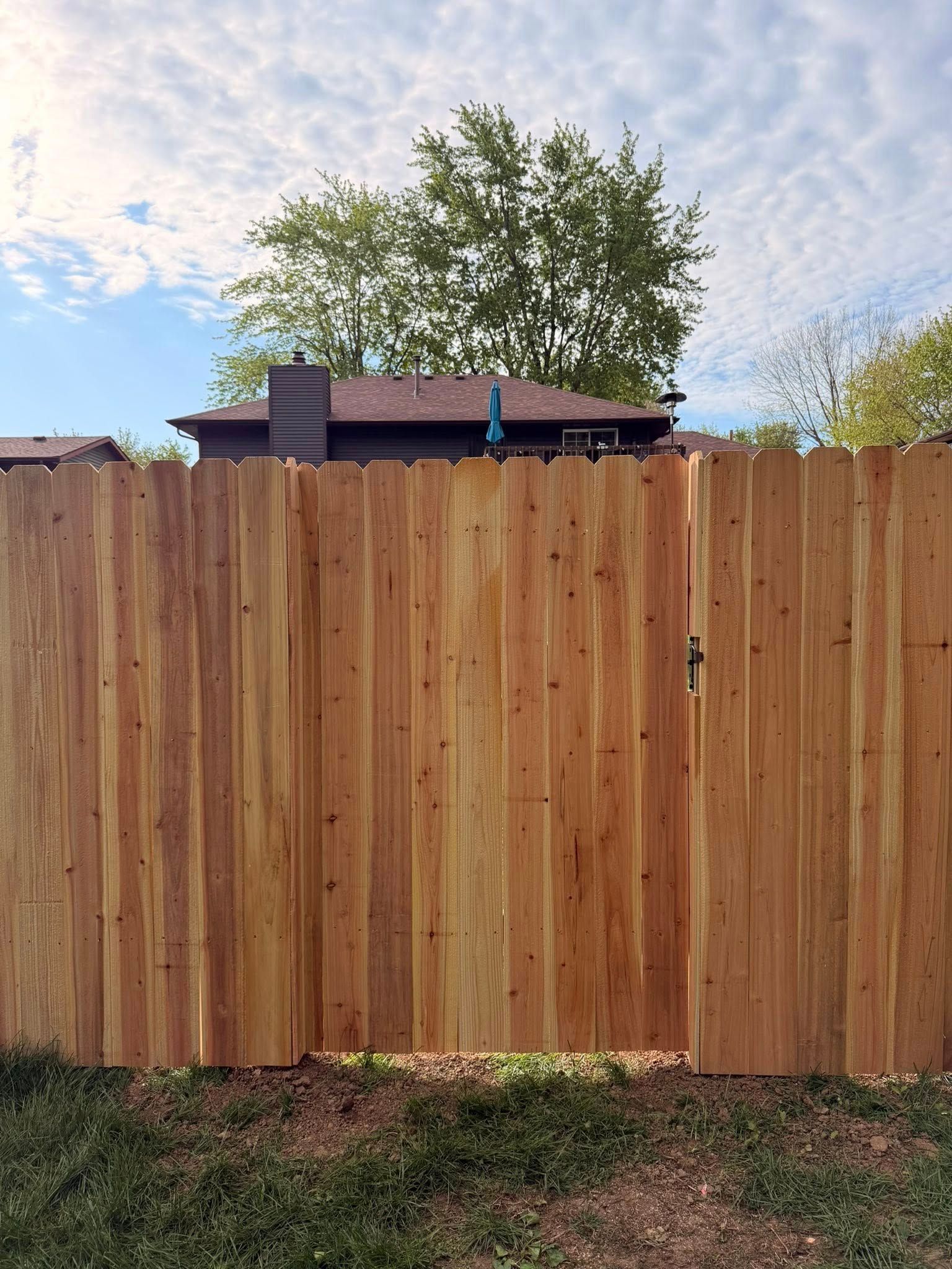 Wooden privacy fence with a tree and house roof visible above it under a partly cloudy sky