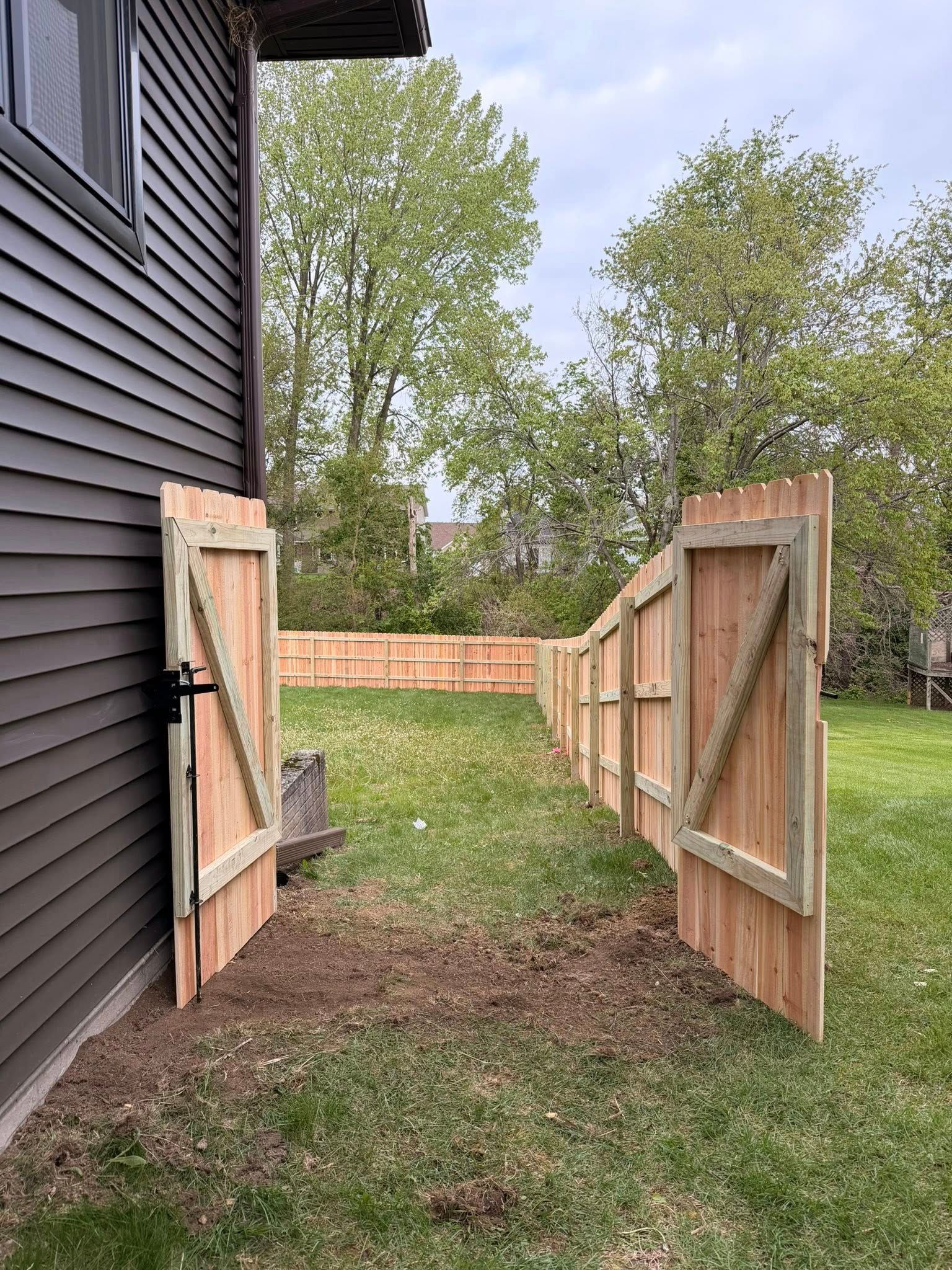 Open wooden gate beside a dark house, leading to a grassy backyard with trees and a brick fence.