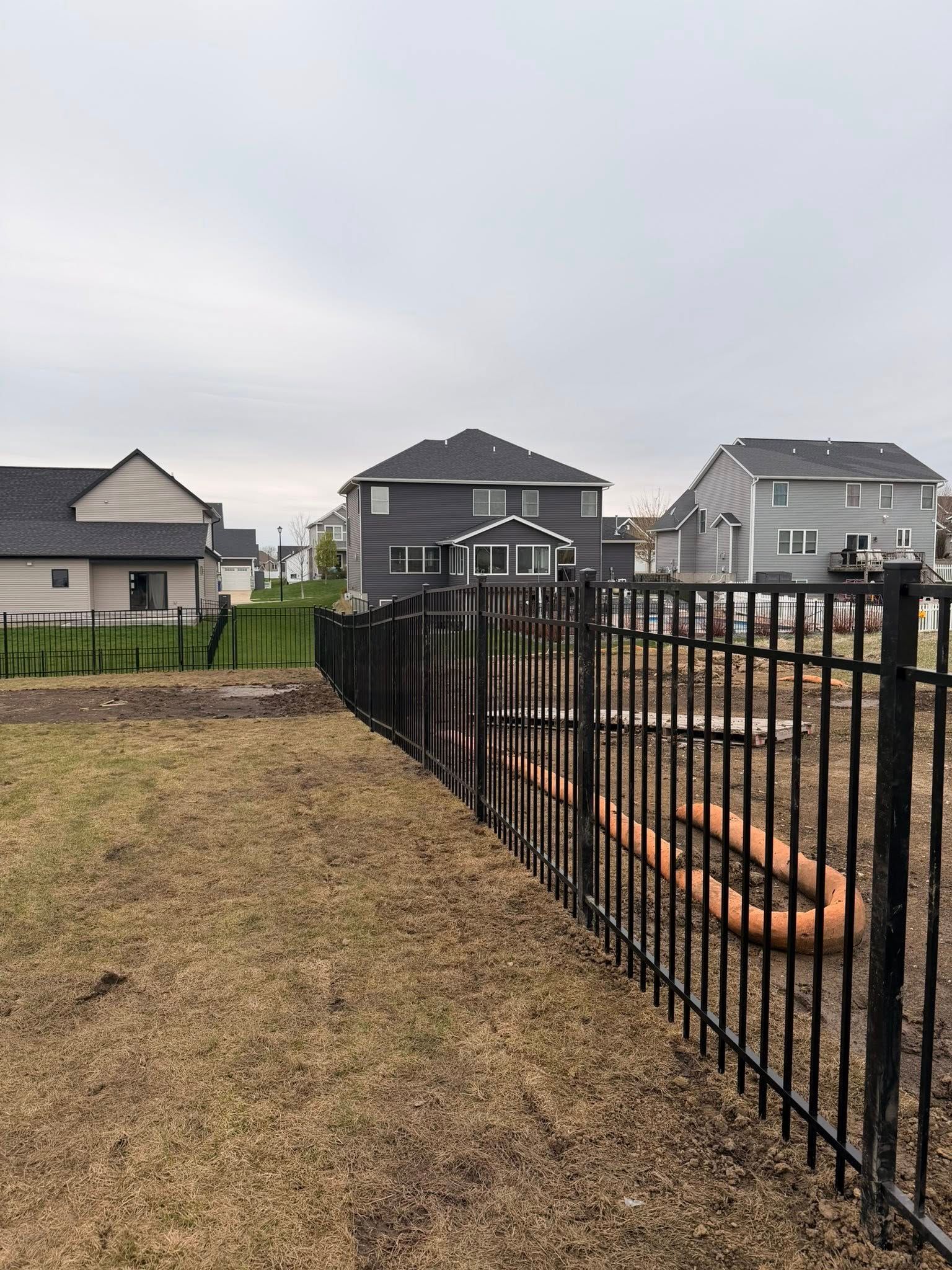 Backyard with black metal fence, bare grass, and gray houses under an overcast sky
