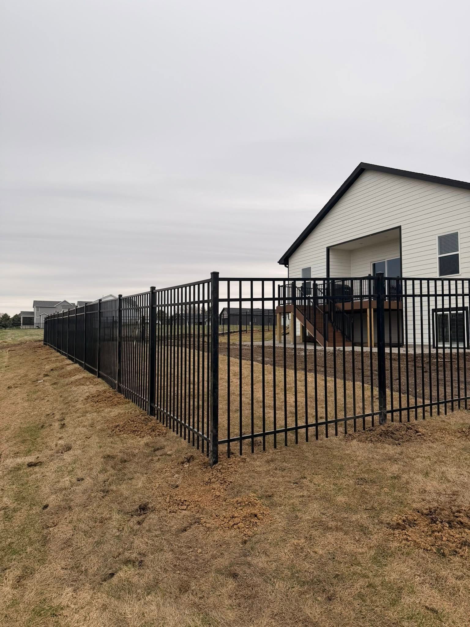 Black metal fence around a beige house on a grassy lot under a gray sky