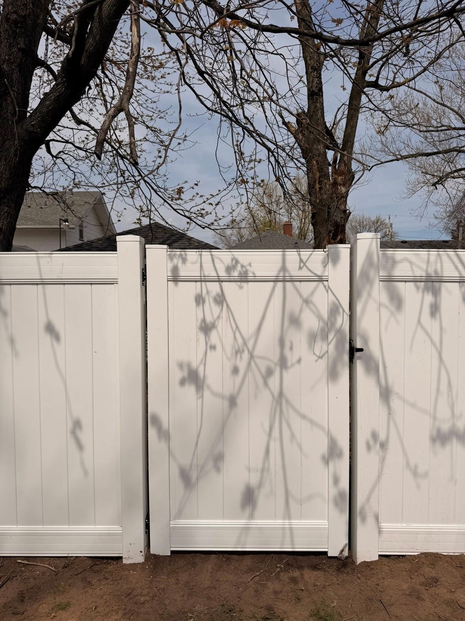 White vinyl fence with tree shadows, leafless branches, and a house roof in the background.
