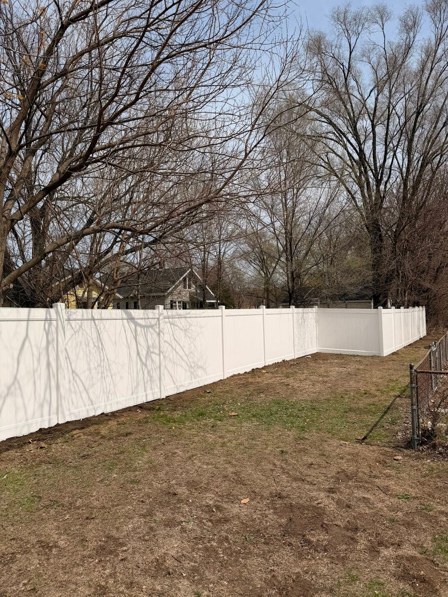 White fence along a leafless backyard with a grassy slope and trees in the background