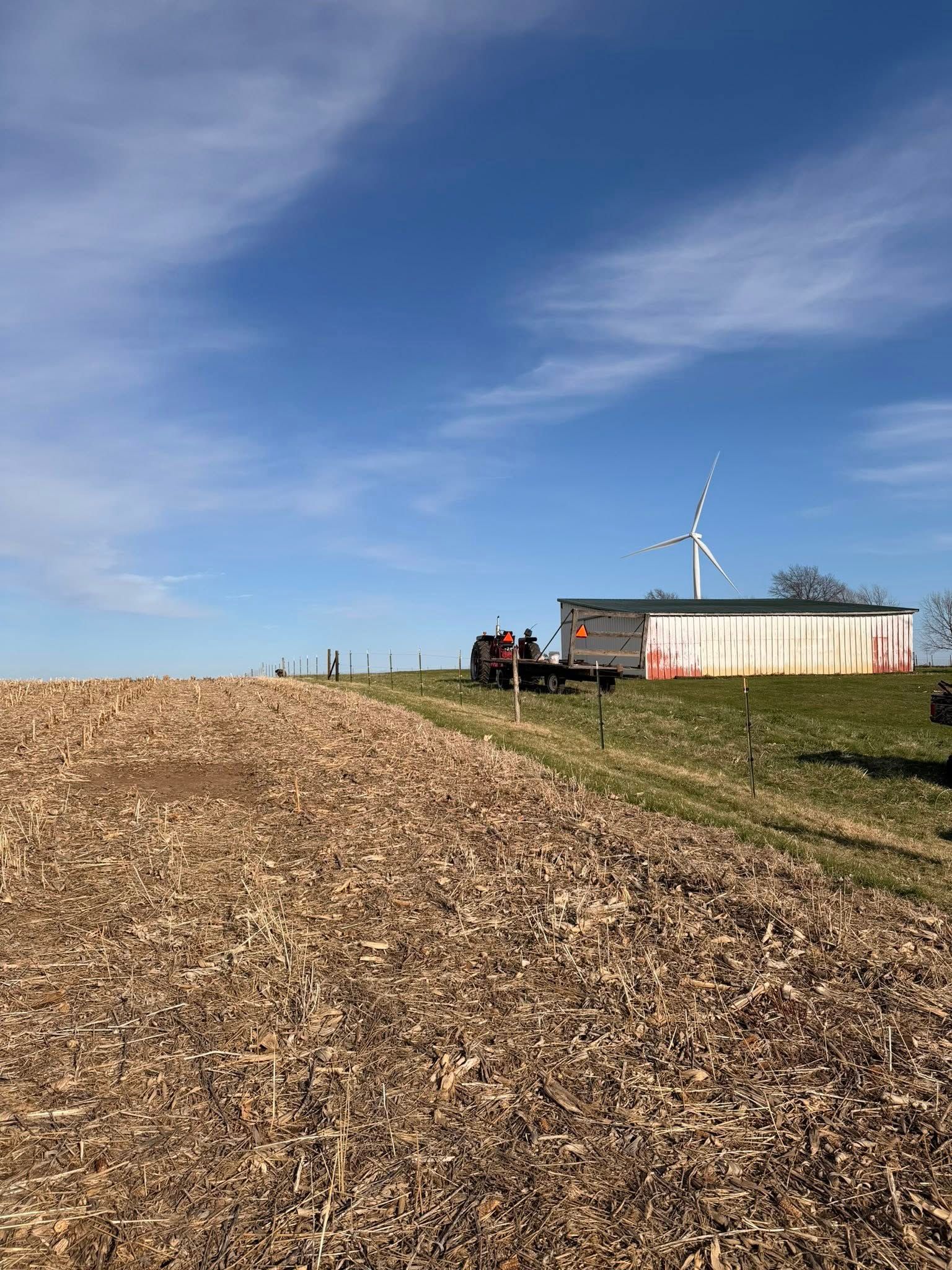 Wide field with a woodchip pile, a small building, and a wind turbine under a blue sky.
