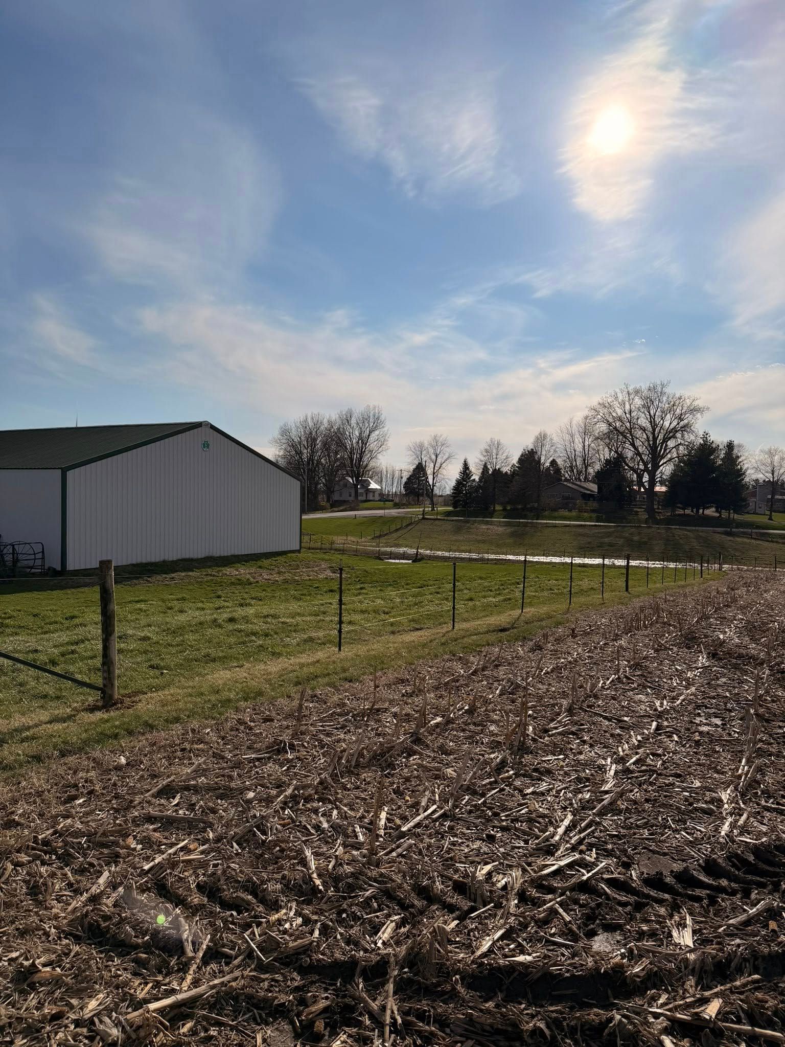 Rural field with a white shed, wire fence, and cloudy blue sky over leaf-covered ground