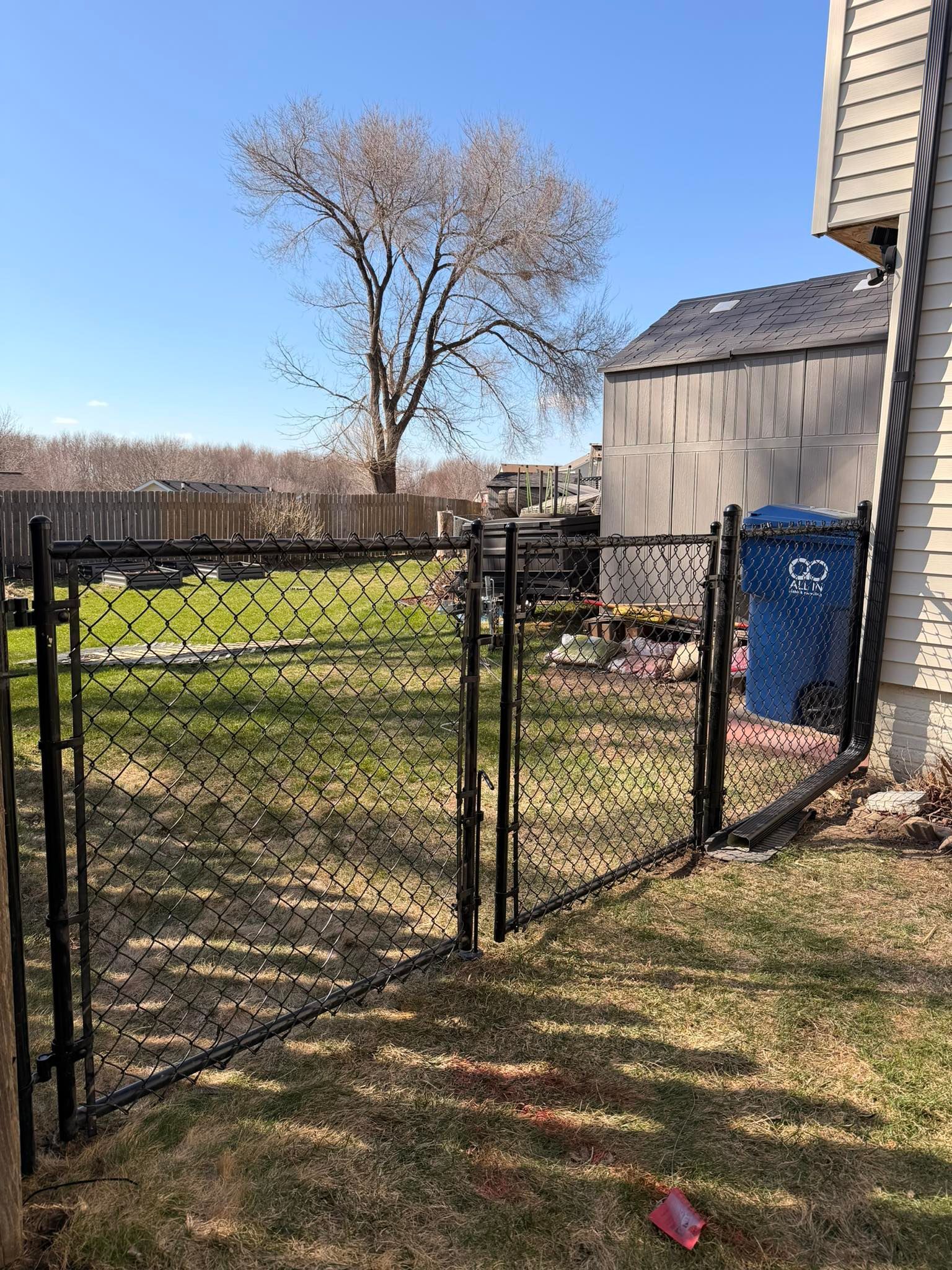 Open black metal gate in a backyard with grass, a tree, and a shed beside a house.