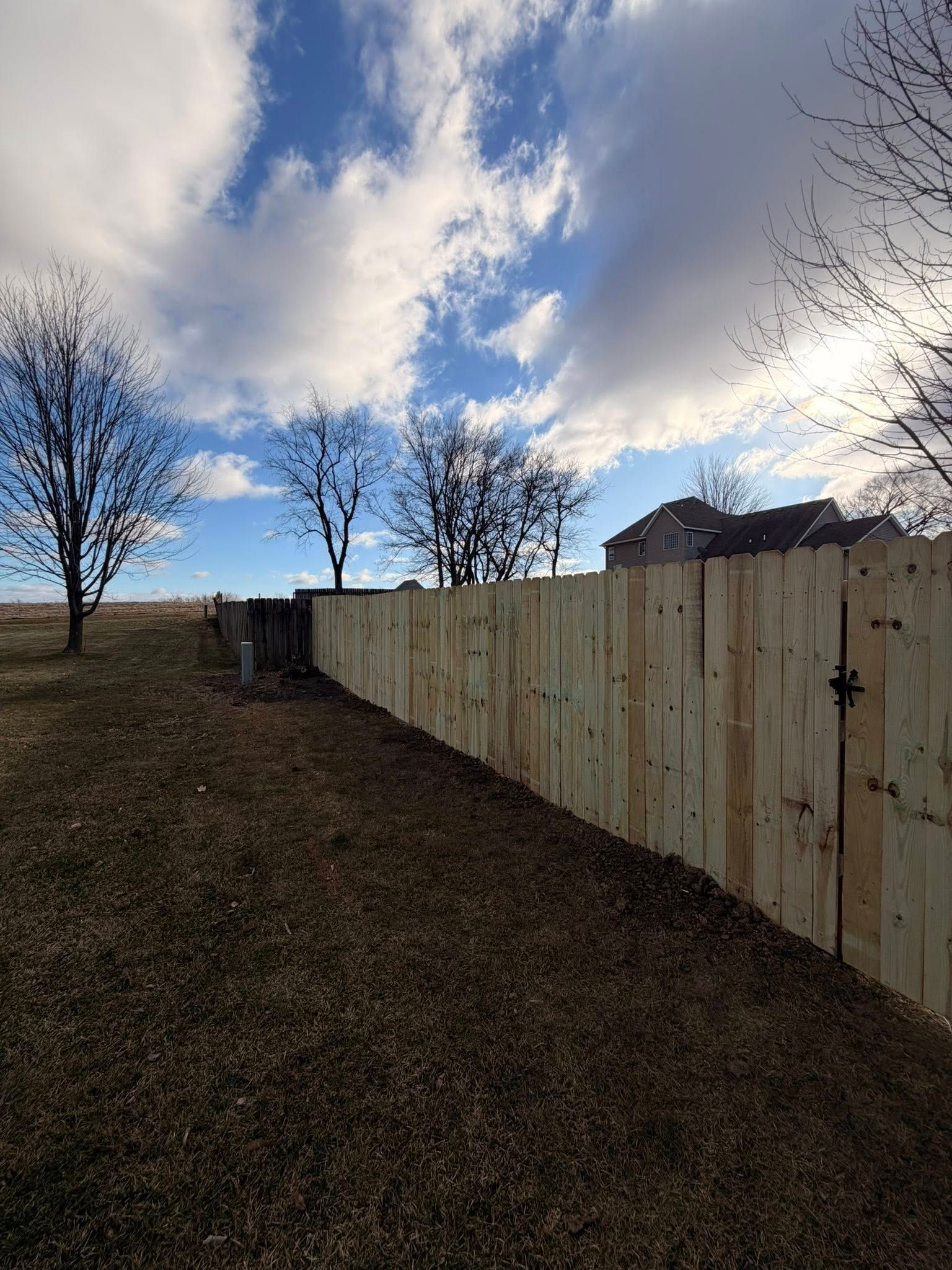 Wooden fence along a grassy slope under a cloudy blue sky with bare trees and houses in the distance