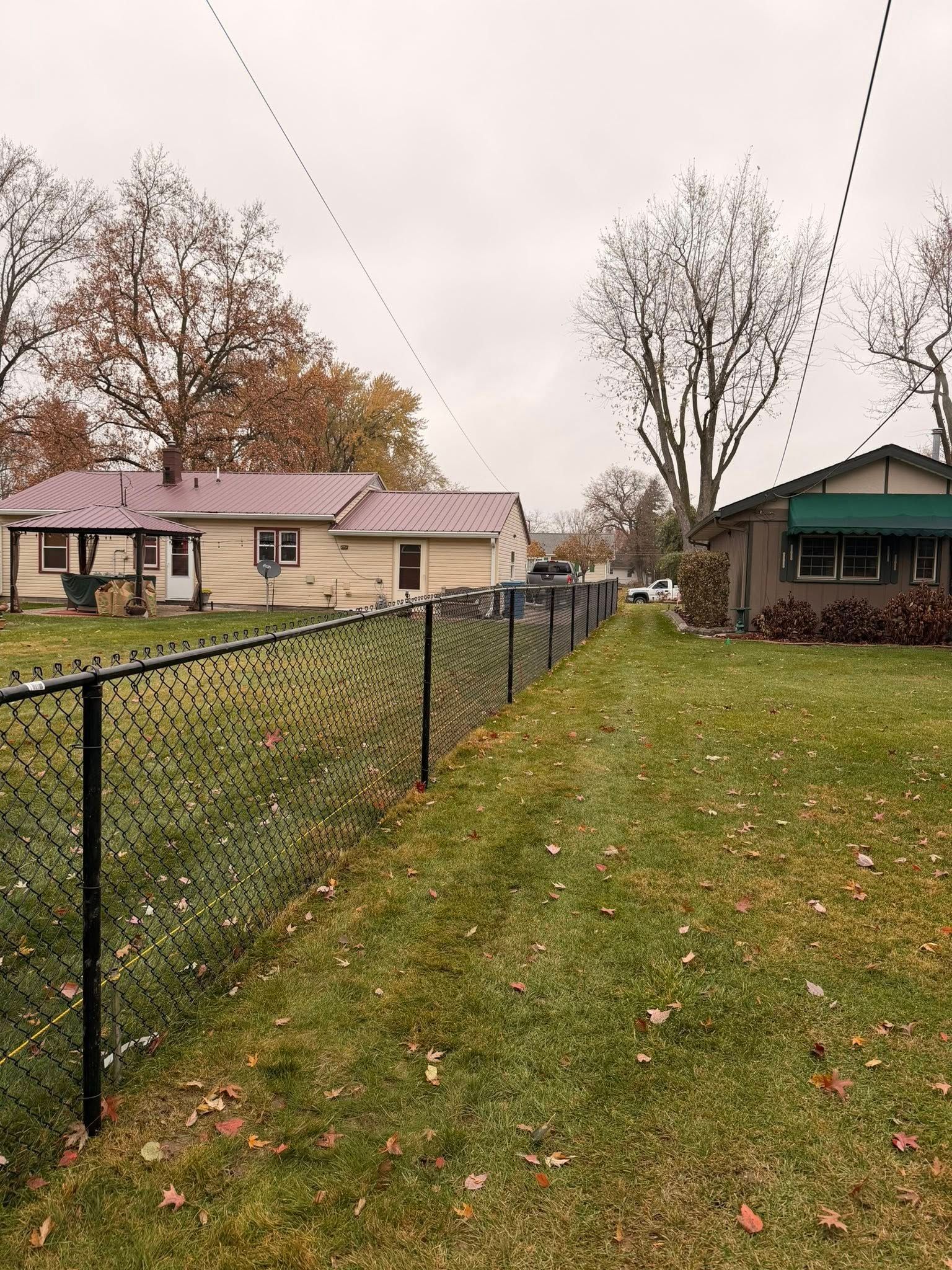 Backyard with chain-link fence, grassy lawn, and two houses under cloudy autumn trees.