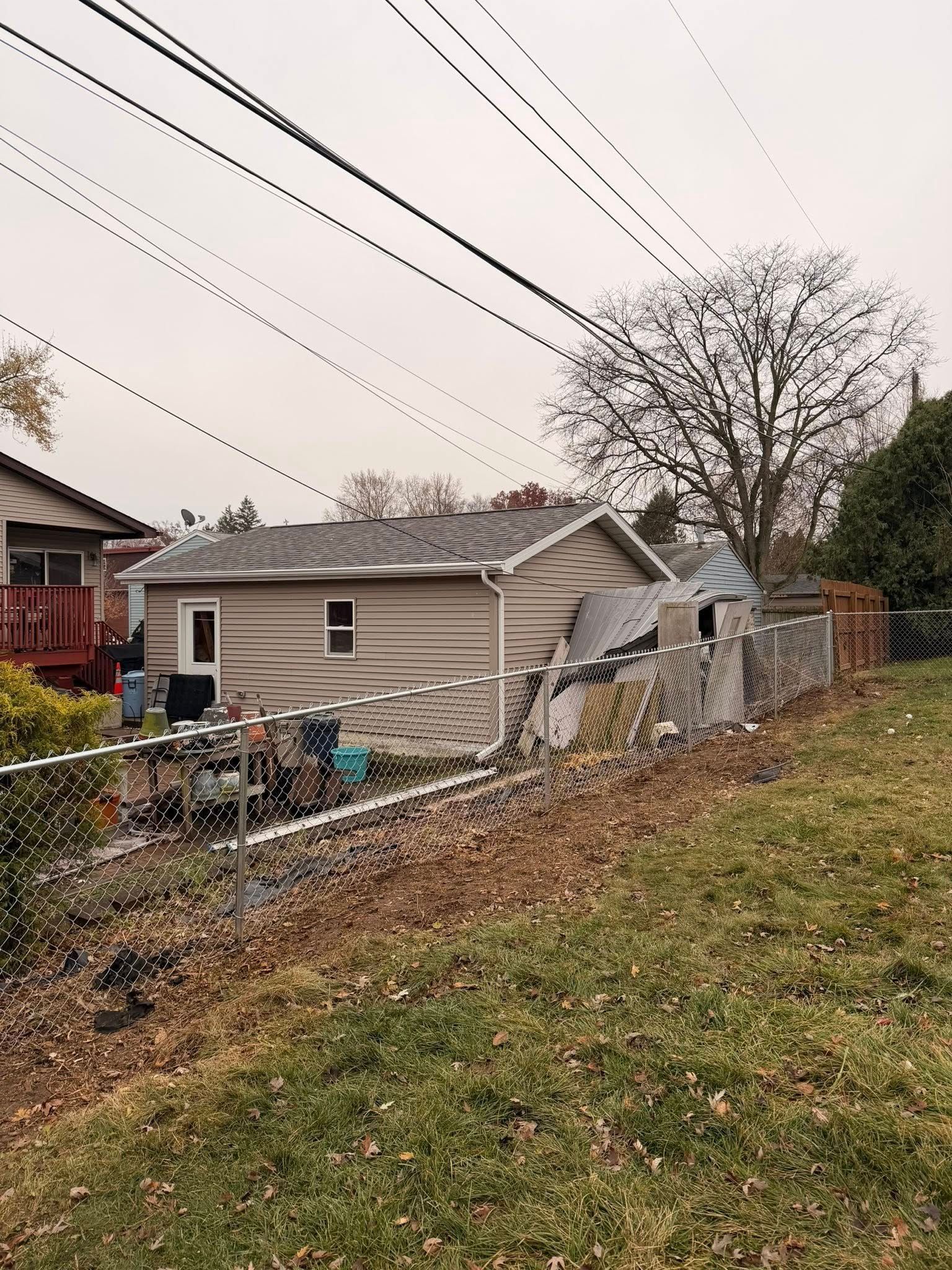 Small beige house with a collapsed fence and debris in a yard under overcast skies.