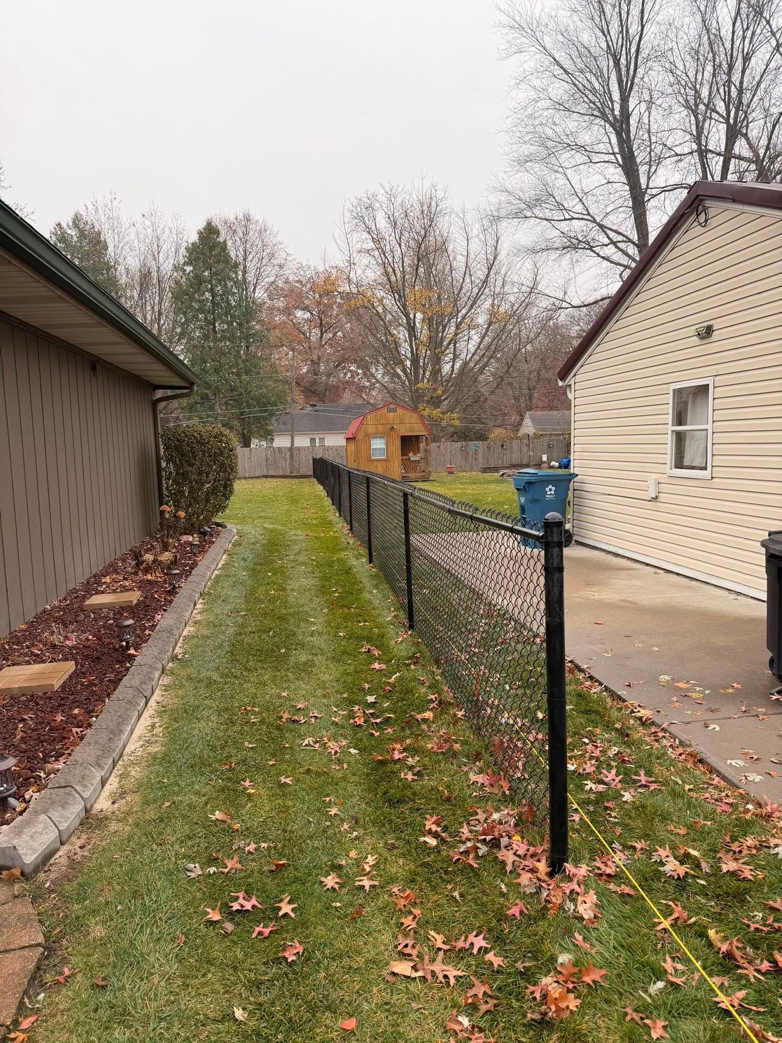 Narrow grassy side yard between two buildings with a black fence and fallen autumn leaves.
