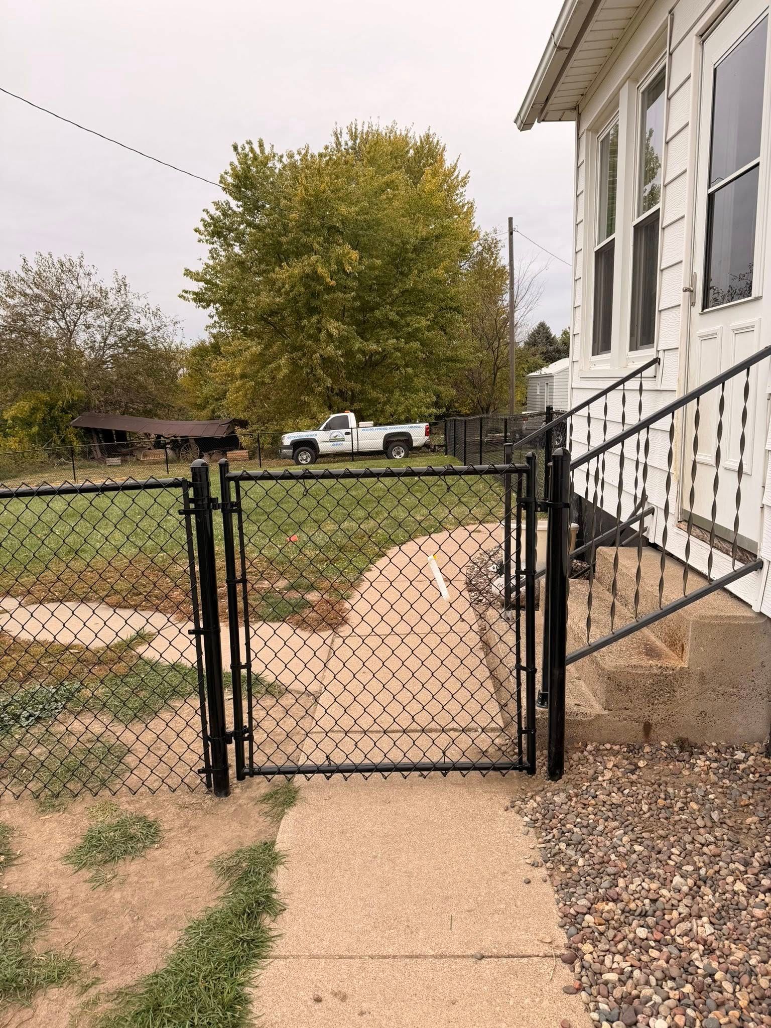 Open gate in chain-link fence beside a white house, facing a grassy yard with a tree and parked pickup truck.