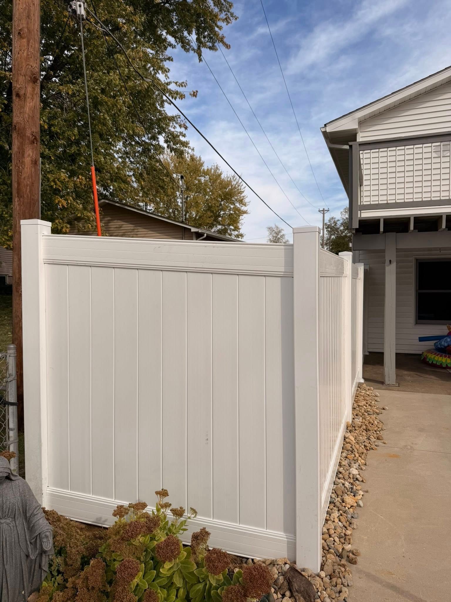 White vinyl fence beside a driveway and house, with trees and a wooden utility pole nearby.