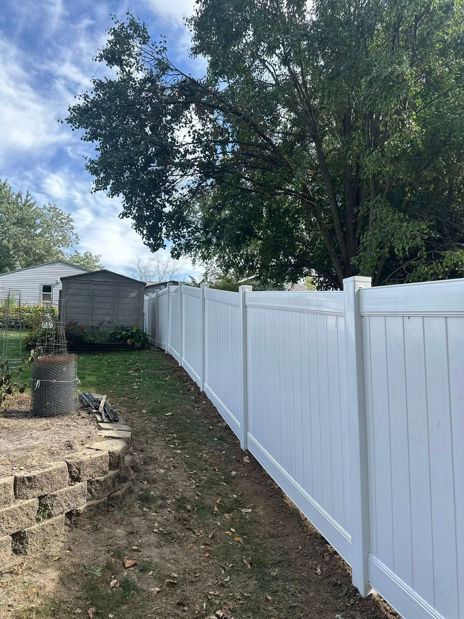White vinyl fence along a grassy backyard beside a large tree and small shed under a cloudy sky