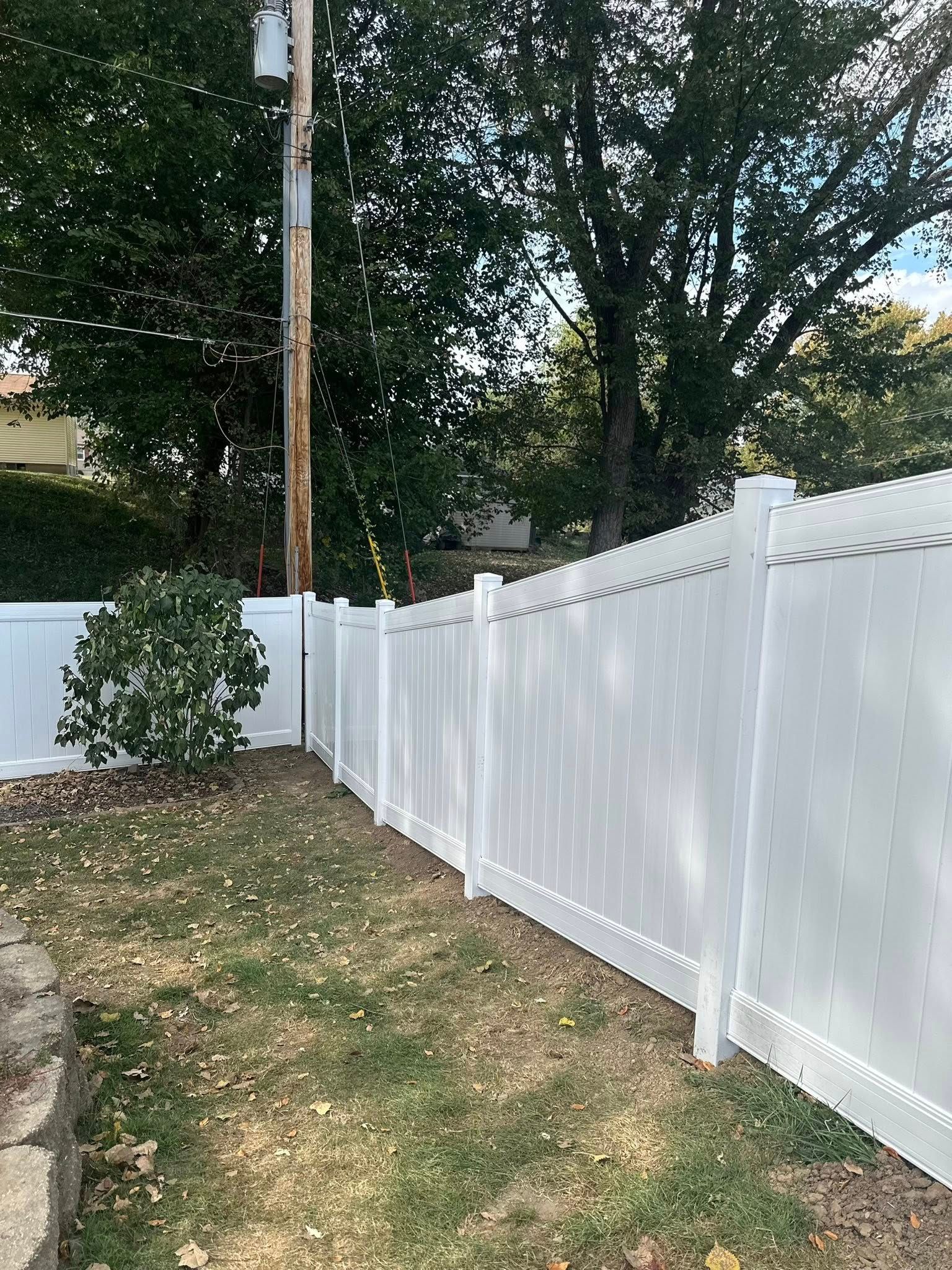 White vinyl fence along a grassy yard with trees and a wooden utility pole behind it.