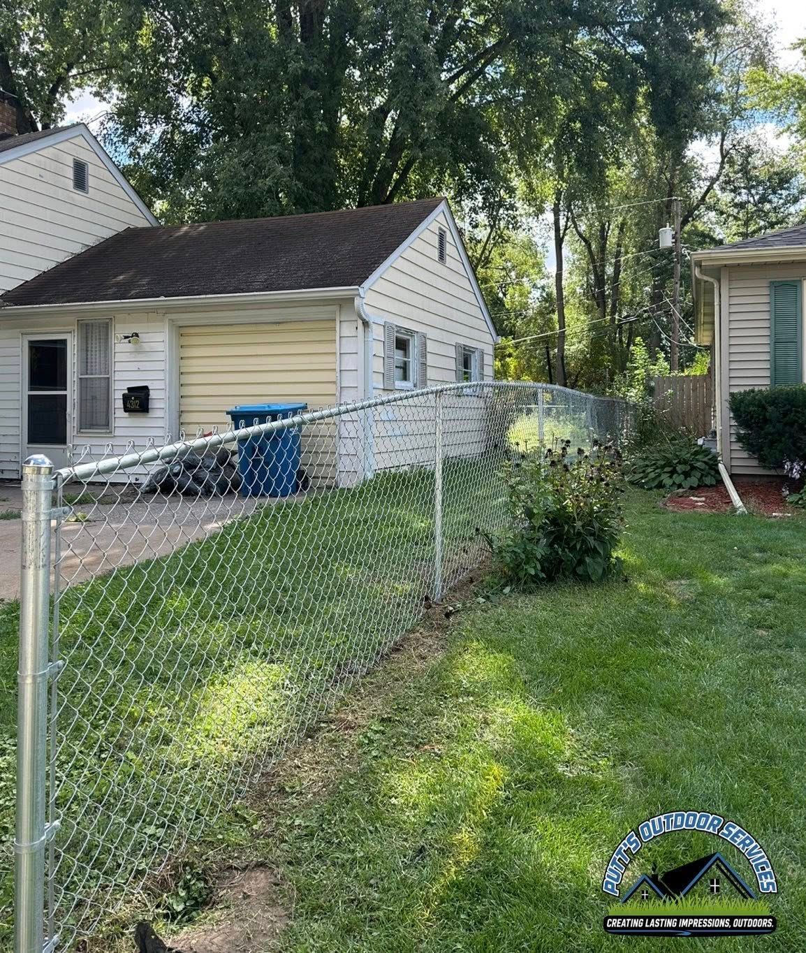 Chain-link fence in a grassy backyard beside small beige houses and tall trees on a sunny day