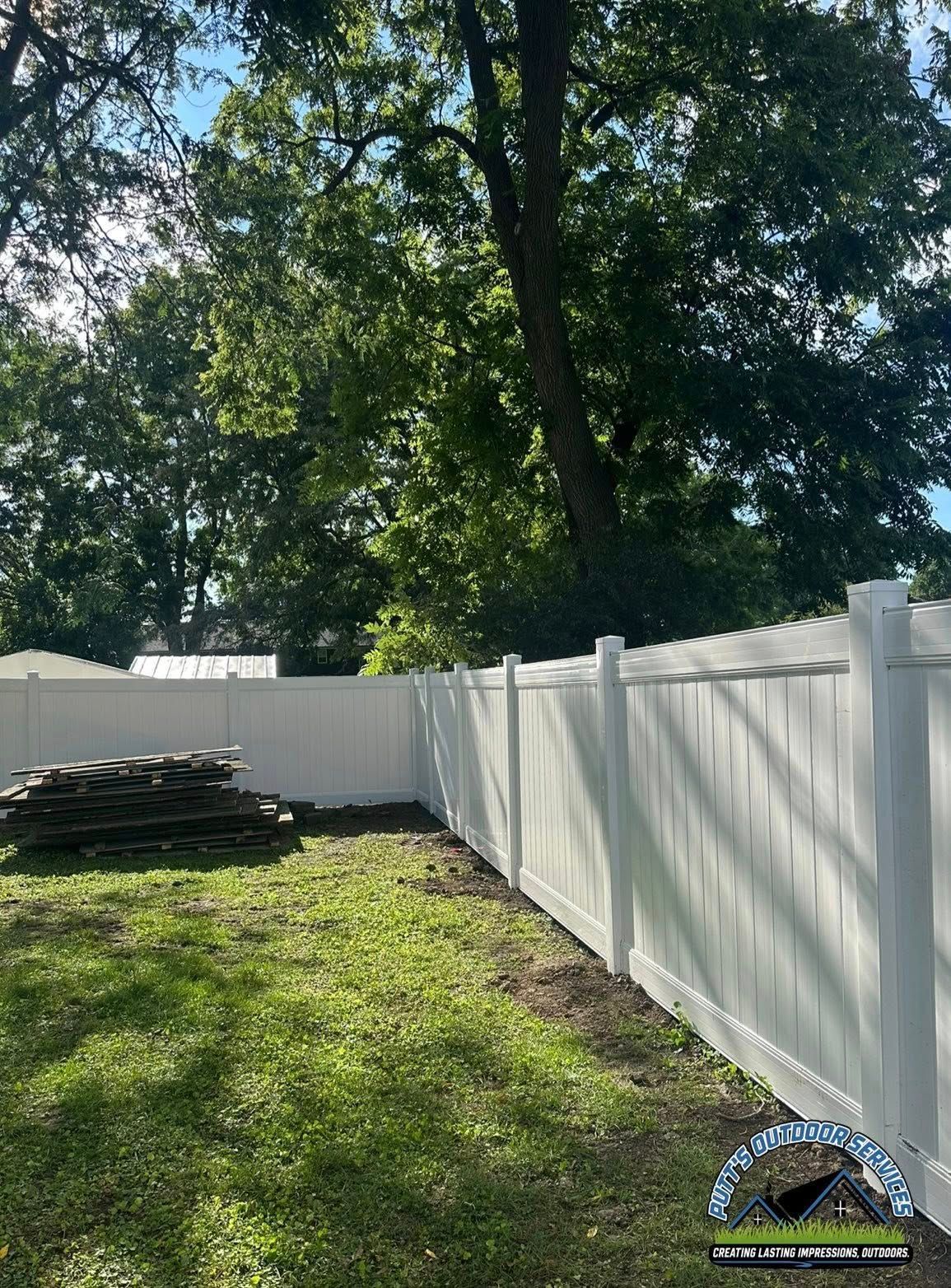 Backyard with white vinyl fence, green grass, tall trees, and stacked materials near the fence.