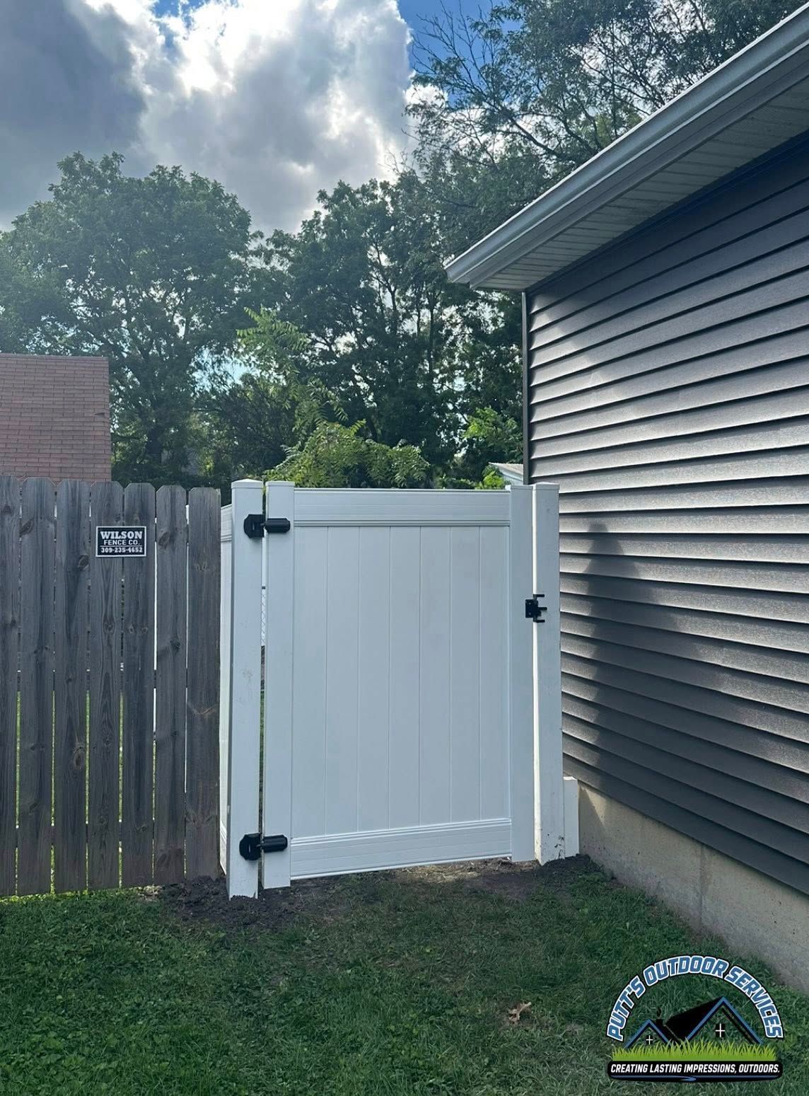White backyard gate between a wooden fence and house siding under a cloudy sky