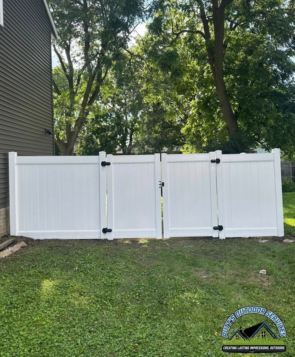 White vinyl fence panels across a grassy yard beside a house, with trees in the background.