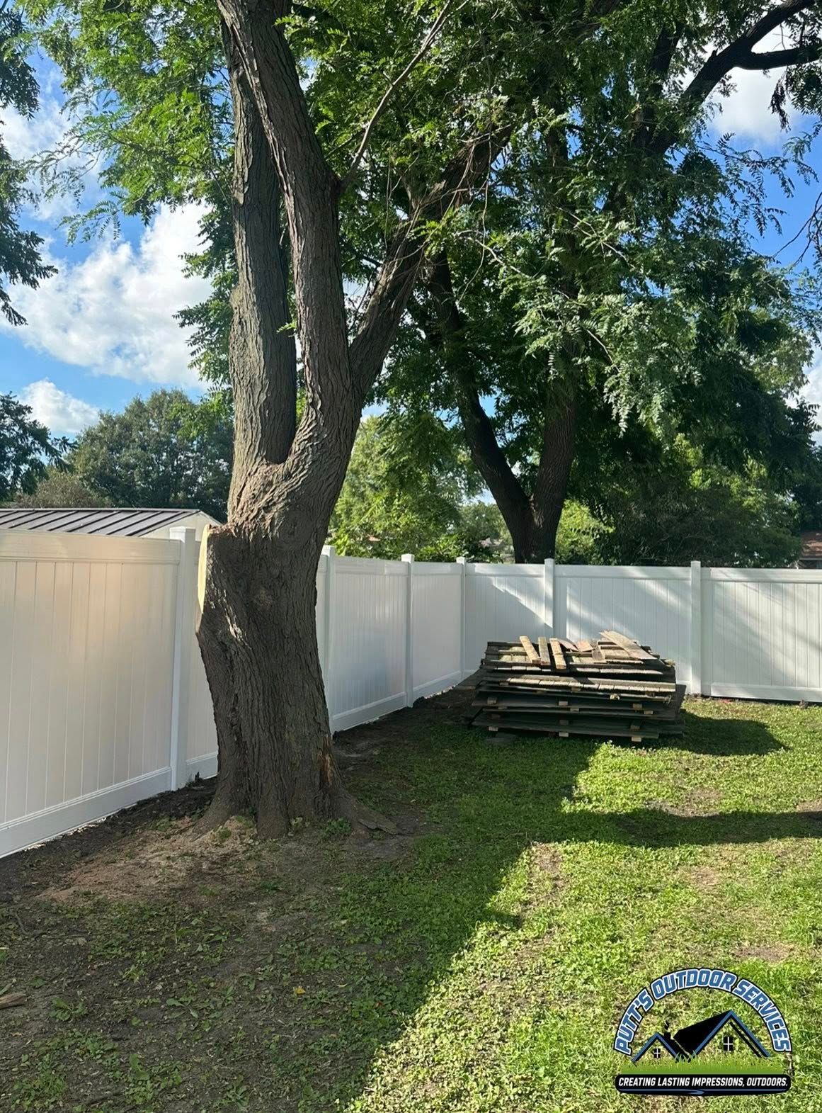 Backyard with large tree, white fence, and stacked wood on grass under a partly cloudy sky