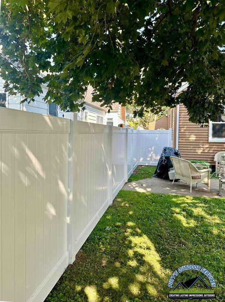 White vinyl fence beside a grassy backyard under a leafy tree, with patio furniture and a small shed area.