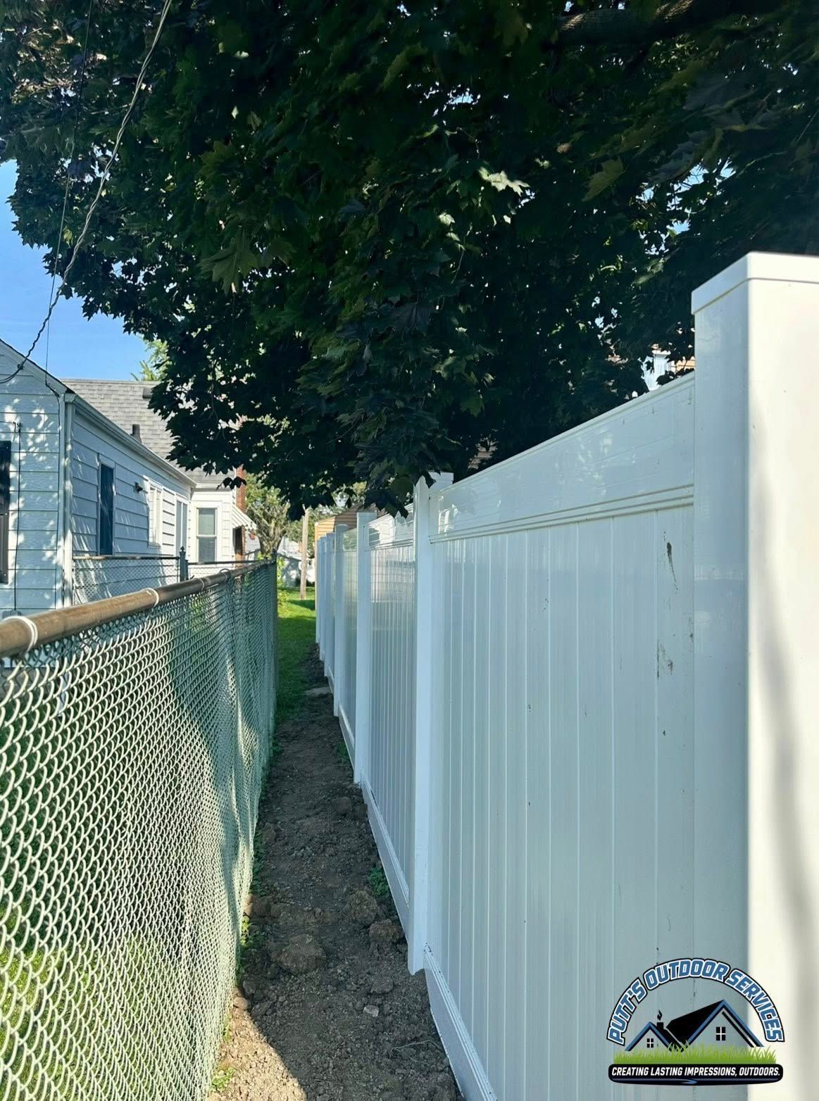 Narrow grassy path between a chain-link fence and white vinyl fence under a large tree.