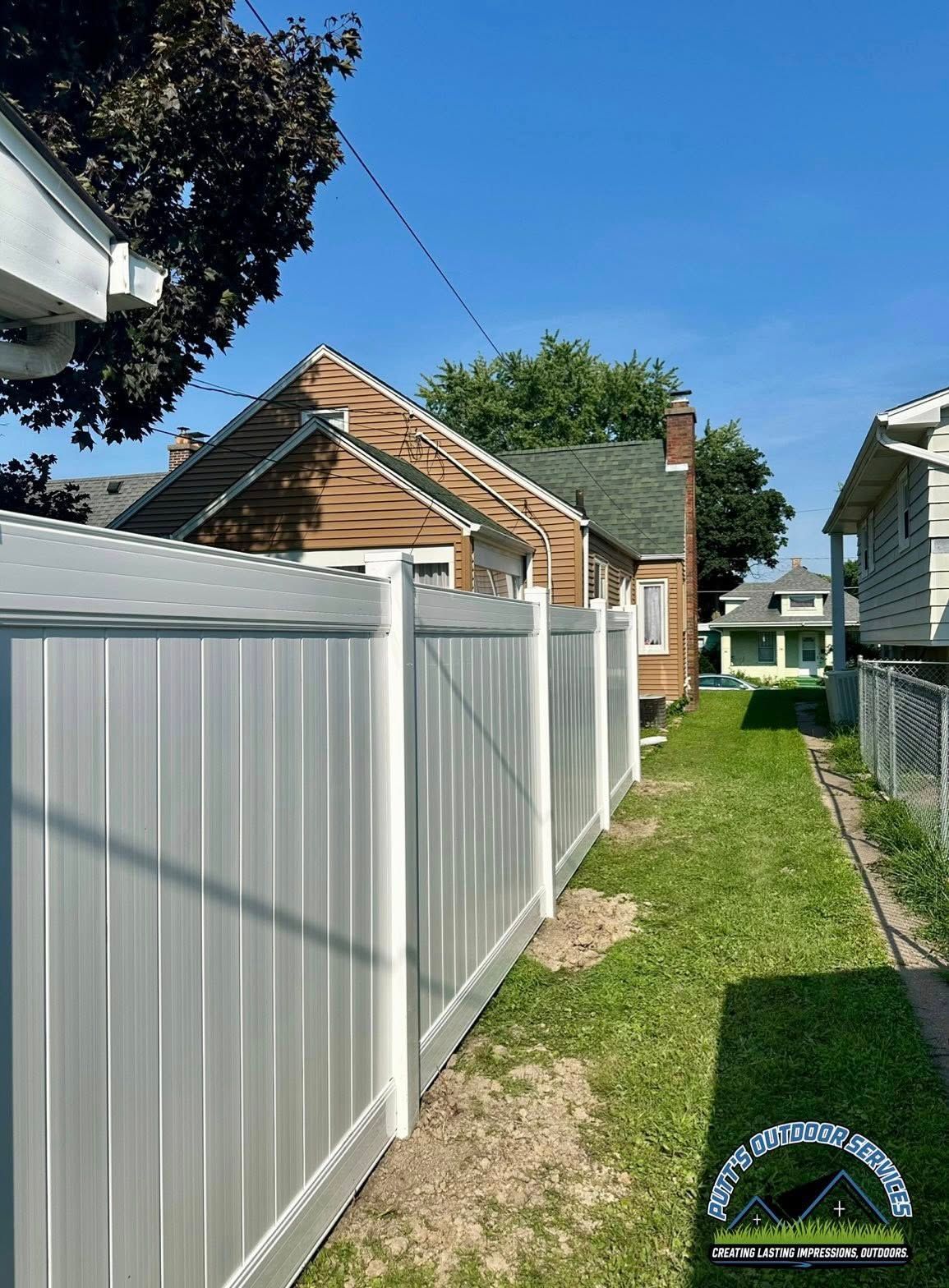 Narrow grassy alley between white vinyl fences beside a house under a blue sky