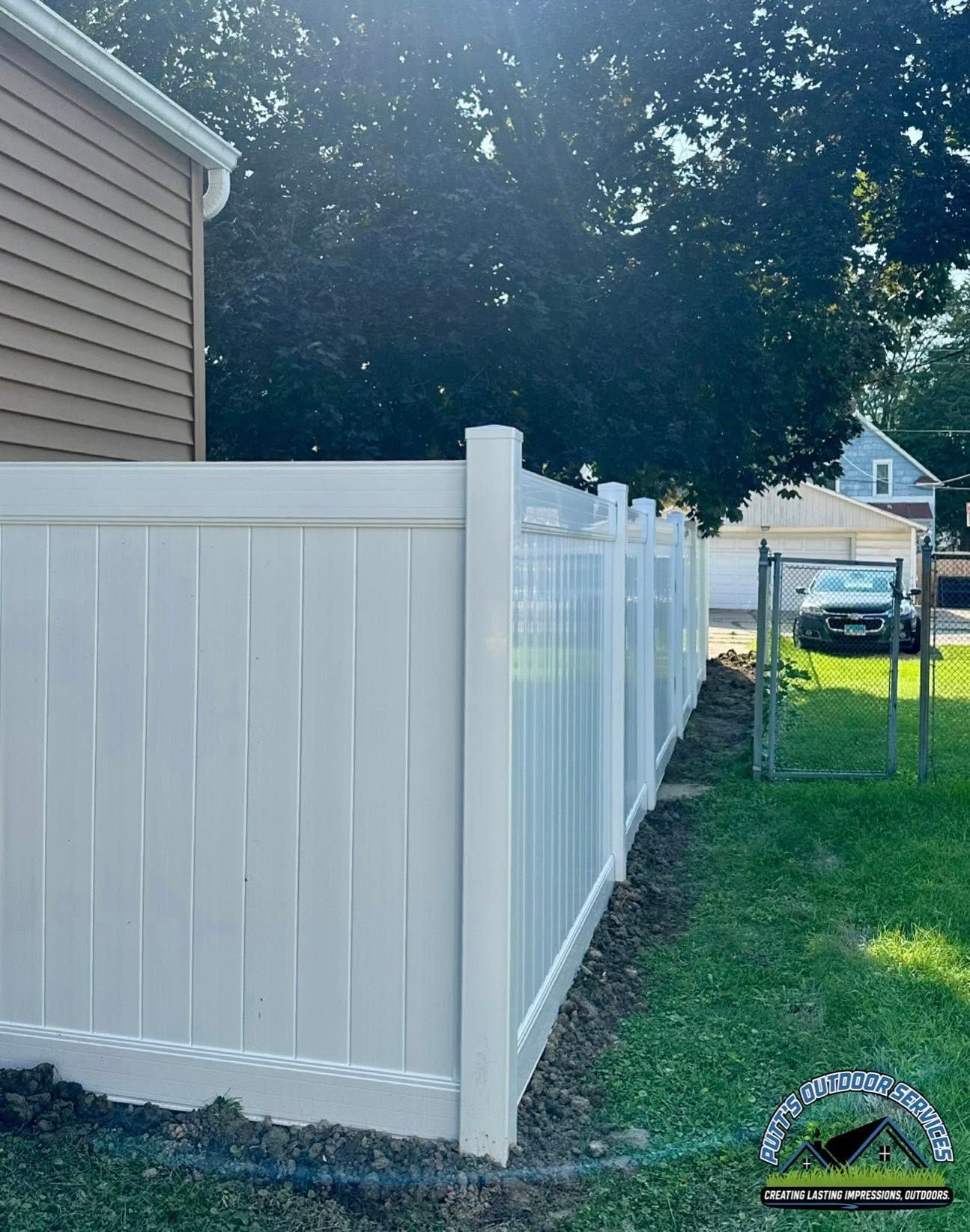 White vinyl fence along a house beside a grassy yard, with trees and a small driveway in the background.