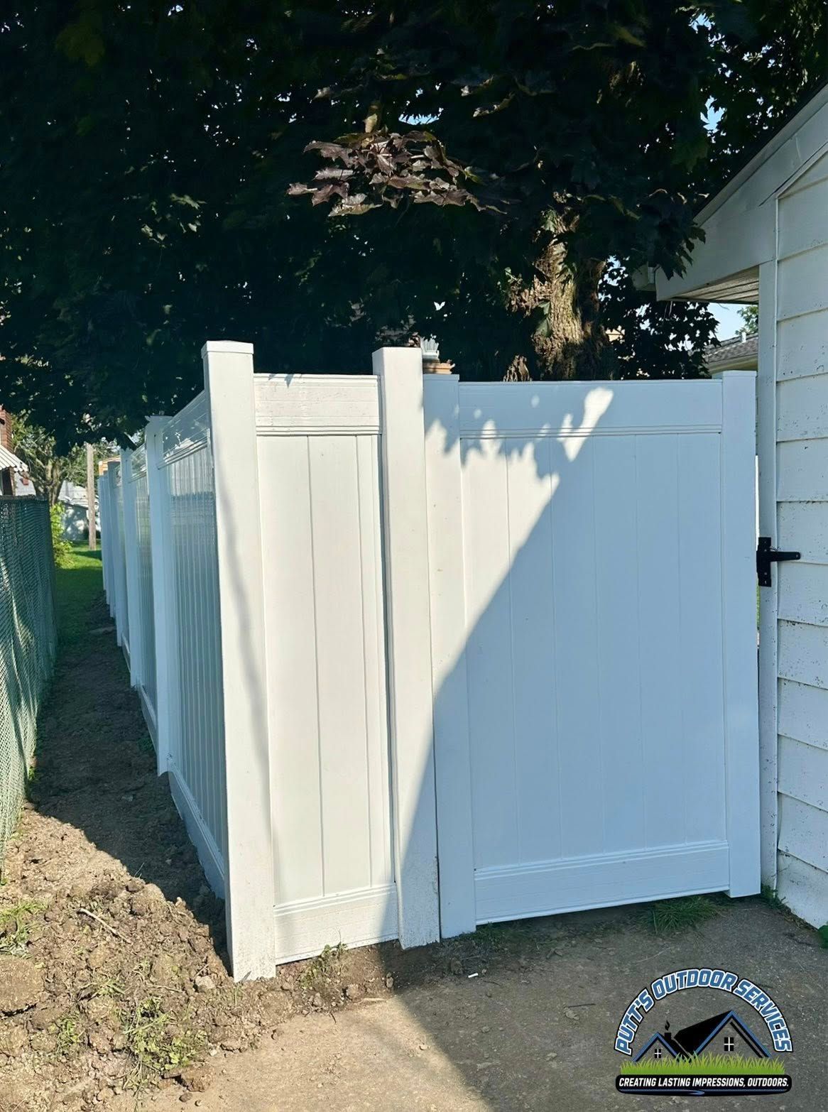 White vinyl fence corner with a gate, beside a gravel path and trees in sunlight.