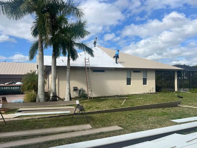 House roof being repaired, with ladder and roof worker near palm trees under a partly cloudy sky