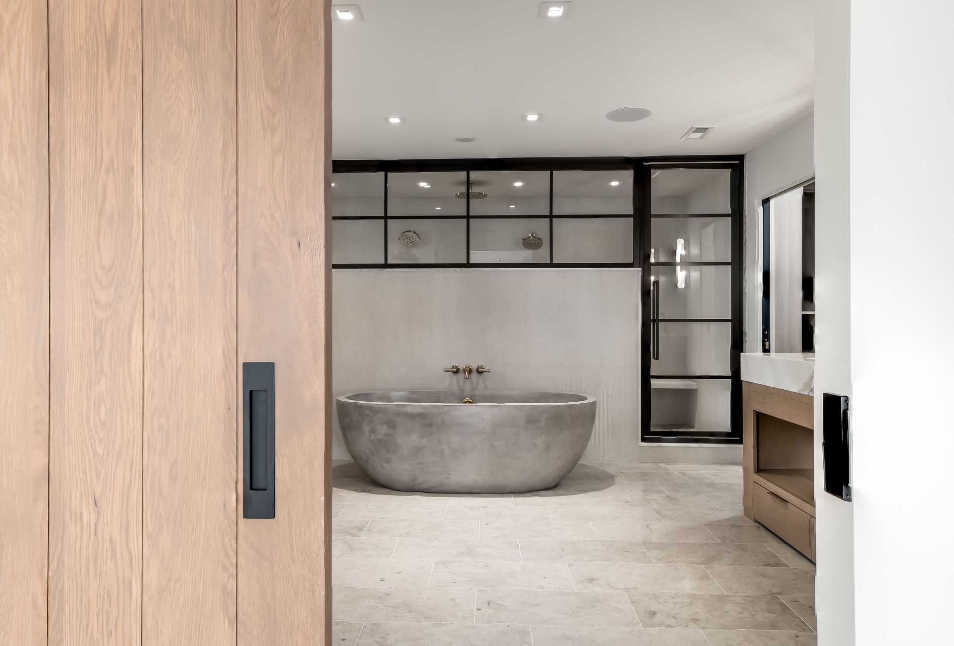 Modern bathroom with a freestanding stone tub, wood vanity, and black-framed glass partition