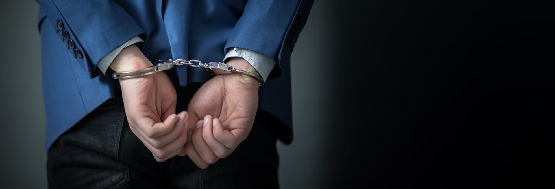 Handcuffed hands behind a person in a blue jacket against a dark background