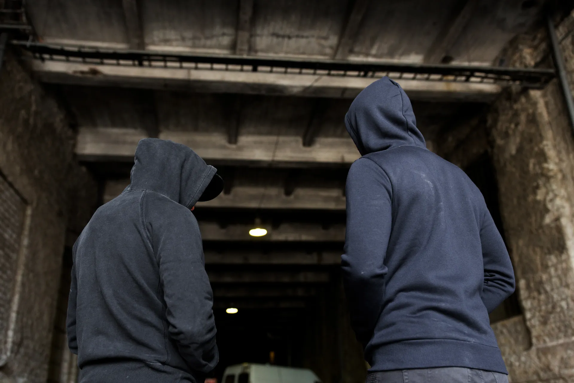 Two hooded people standing at a dark concrete tunnel entrance
