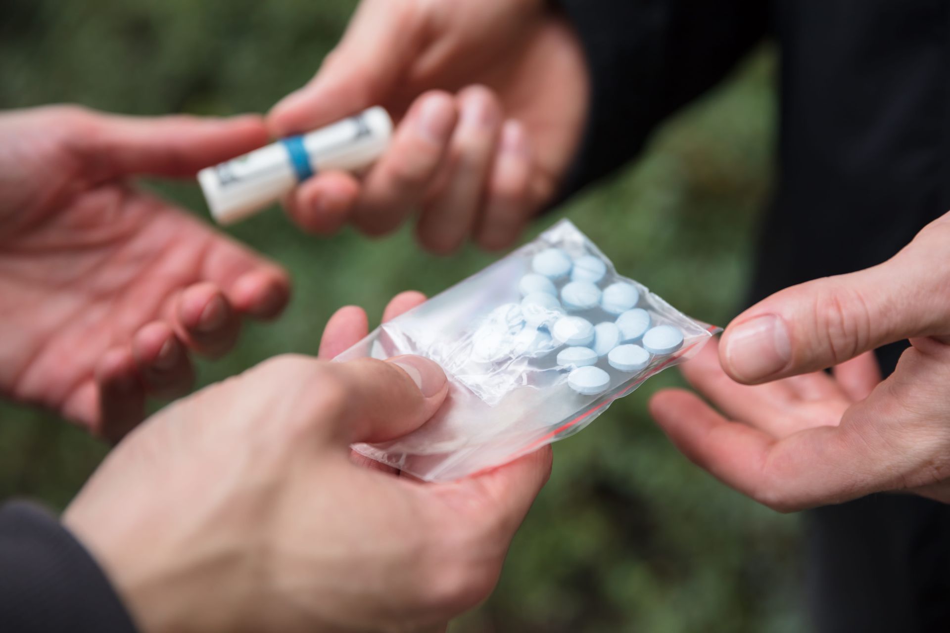 Hands exchange a clear bag of white tablets and a pill bottle outdoors.