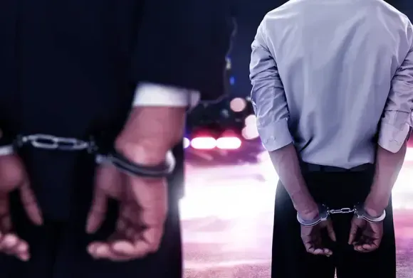 Handcuffed person standing with police officers in a dark scene with flashing lights