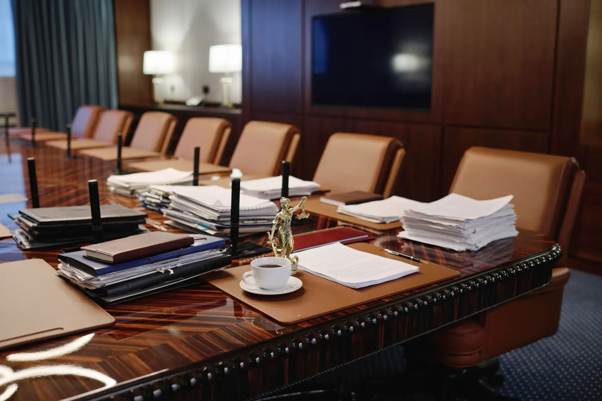 Conference room table with papers, notebooks, pens, and water pitcher beside tan chairs and a wall-mounted TV