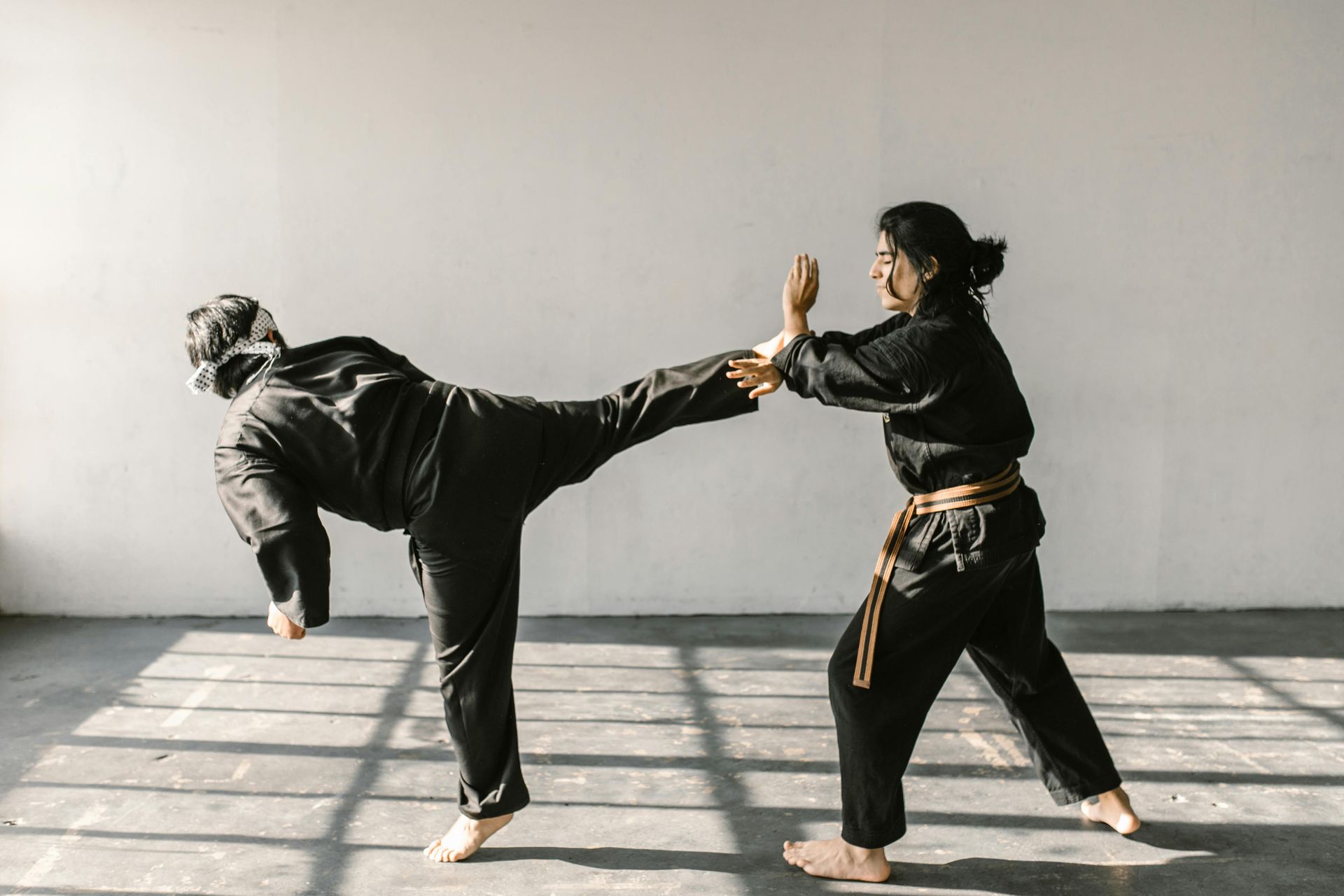 Two martial artists spar in a studio, one kicking while the other blocks with raised hands.