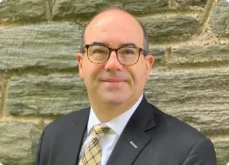A person wearing a dark suit, white shirt, and patterned tie, standing in front of a light gray stone wall.