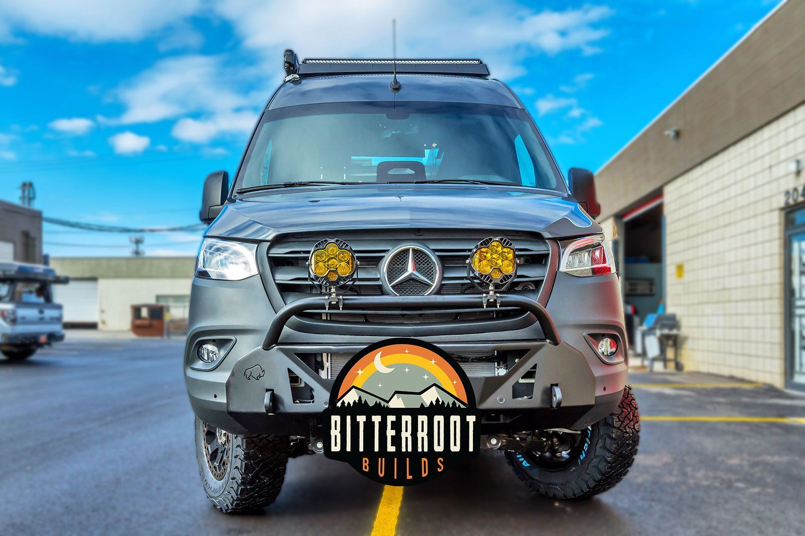 Front view of a gray Mercedes off-road van with yellow lights and Bitterroot Builds plate in a shop lot