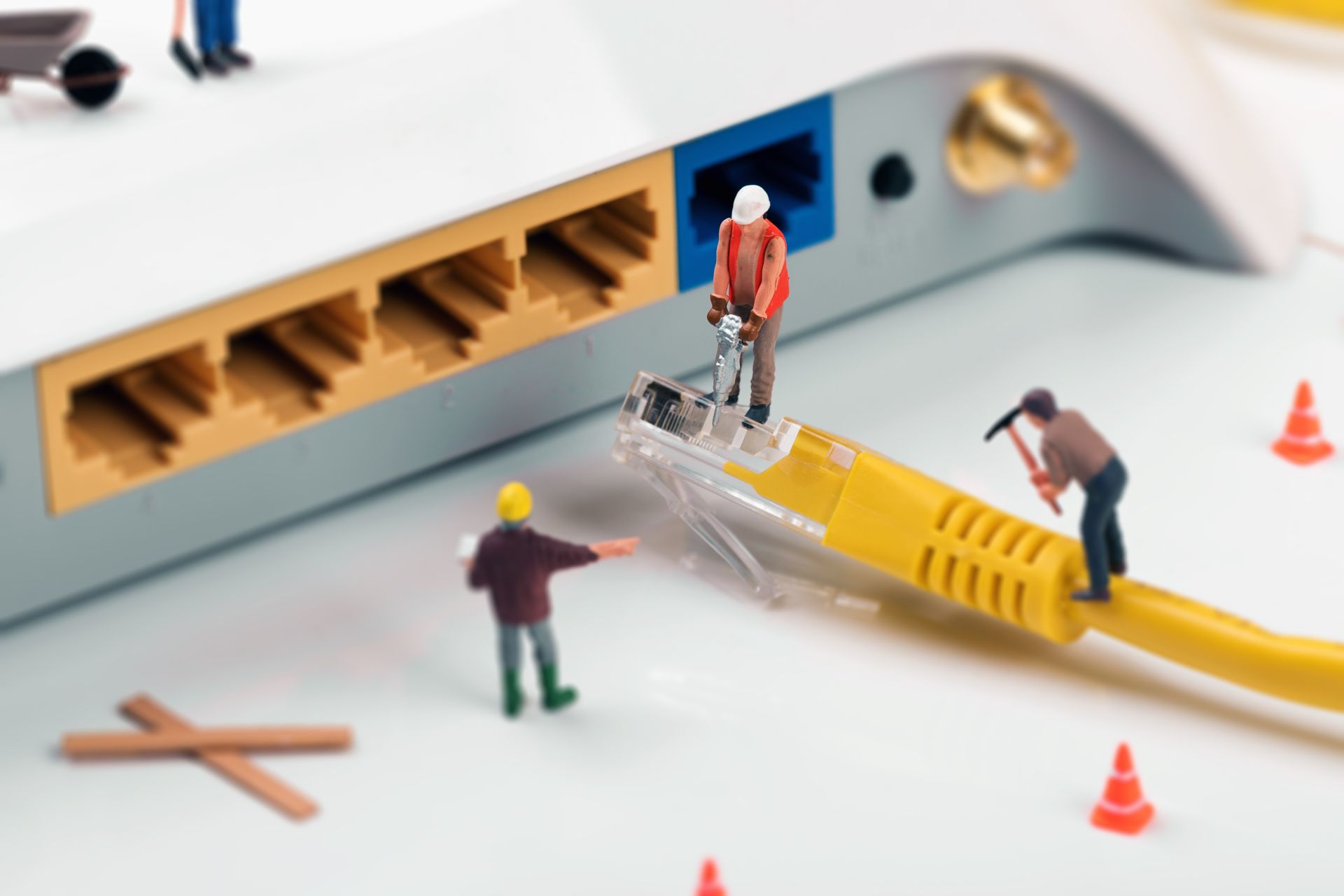 Miniature workers repairing a yellow ethernet cable connected to a router, with orange safety cones.