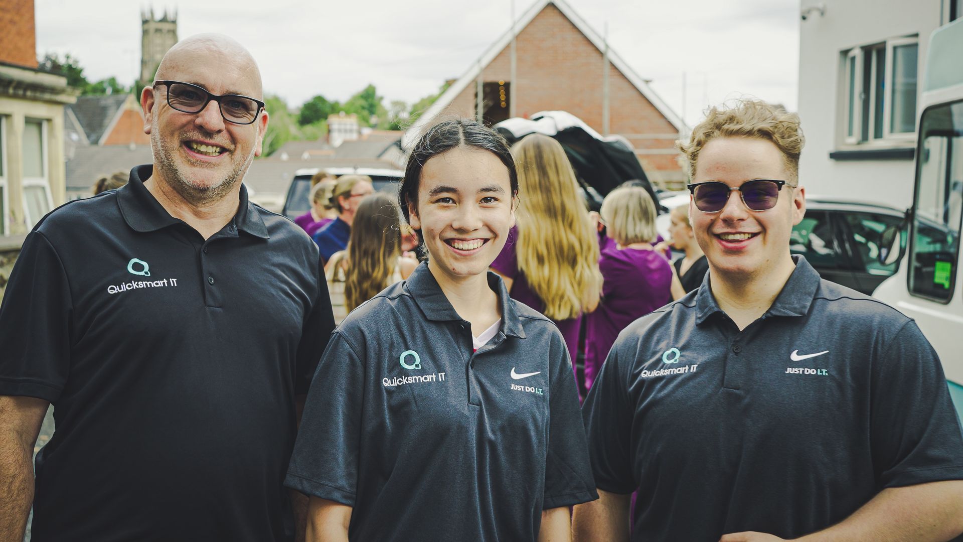 Three smiling people in black polo shirts standing outdoors in front of houses