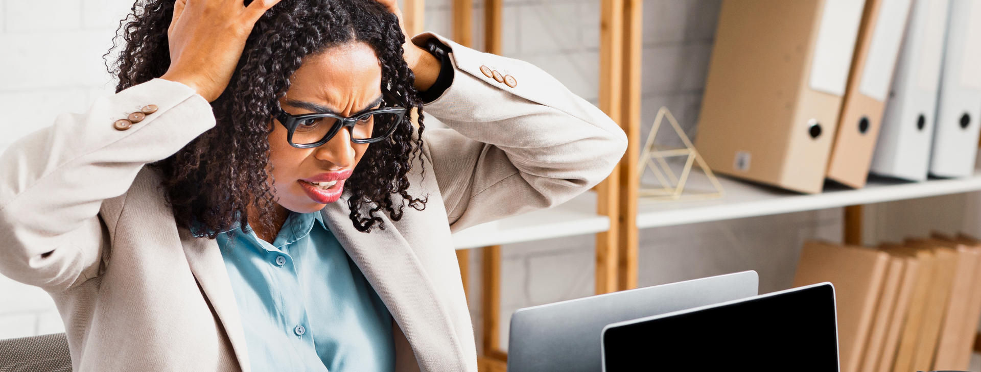 Woman with glasses, stressed at a computer, touching her head in an office setting.