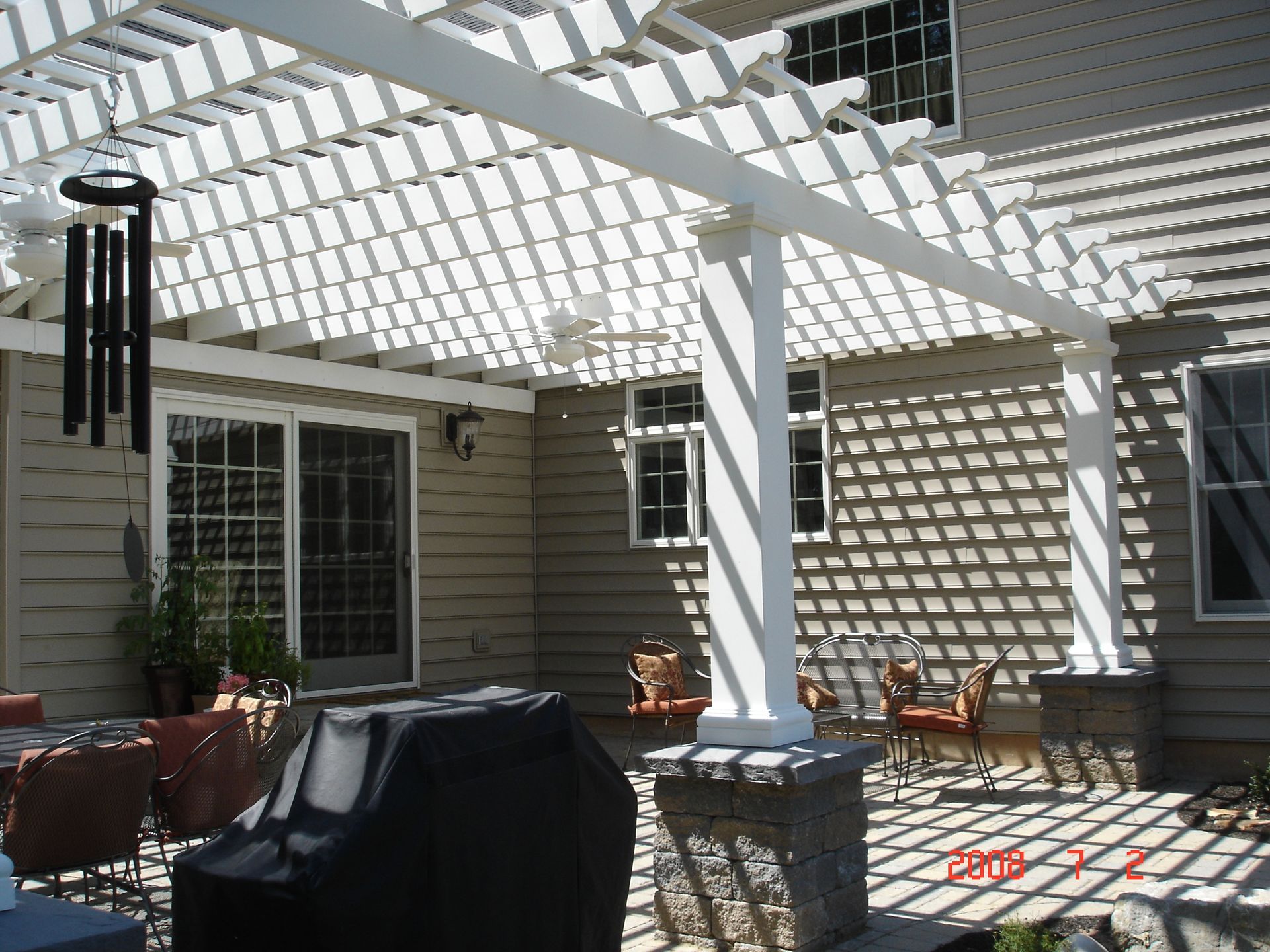 Patio with white pergola casting shadows, beige house siding, and outdoor seating in bright sunlight