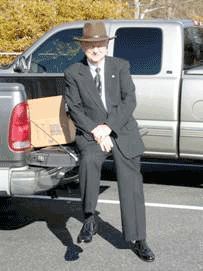 Man in a dark suit and hat leaning against a pickup truck in a parking lot