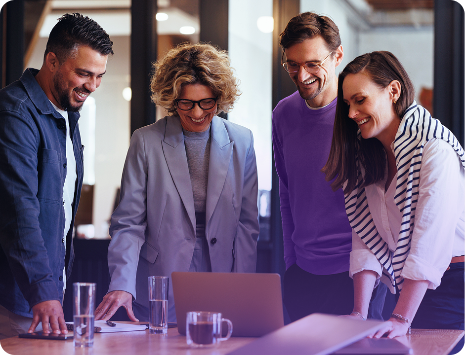 Quatro colegas de trabalho sorrindo, reunidos em volta de um laptop em um escritório, discutindo algo.