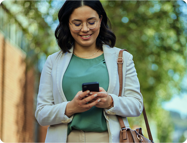 Mulher de óculos sorri enquanto olha para o celular; caminhando ao ar livre.