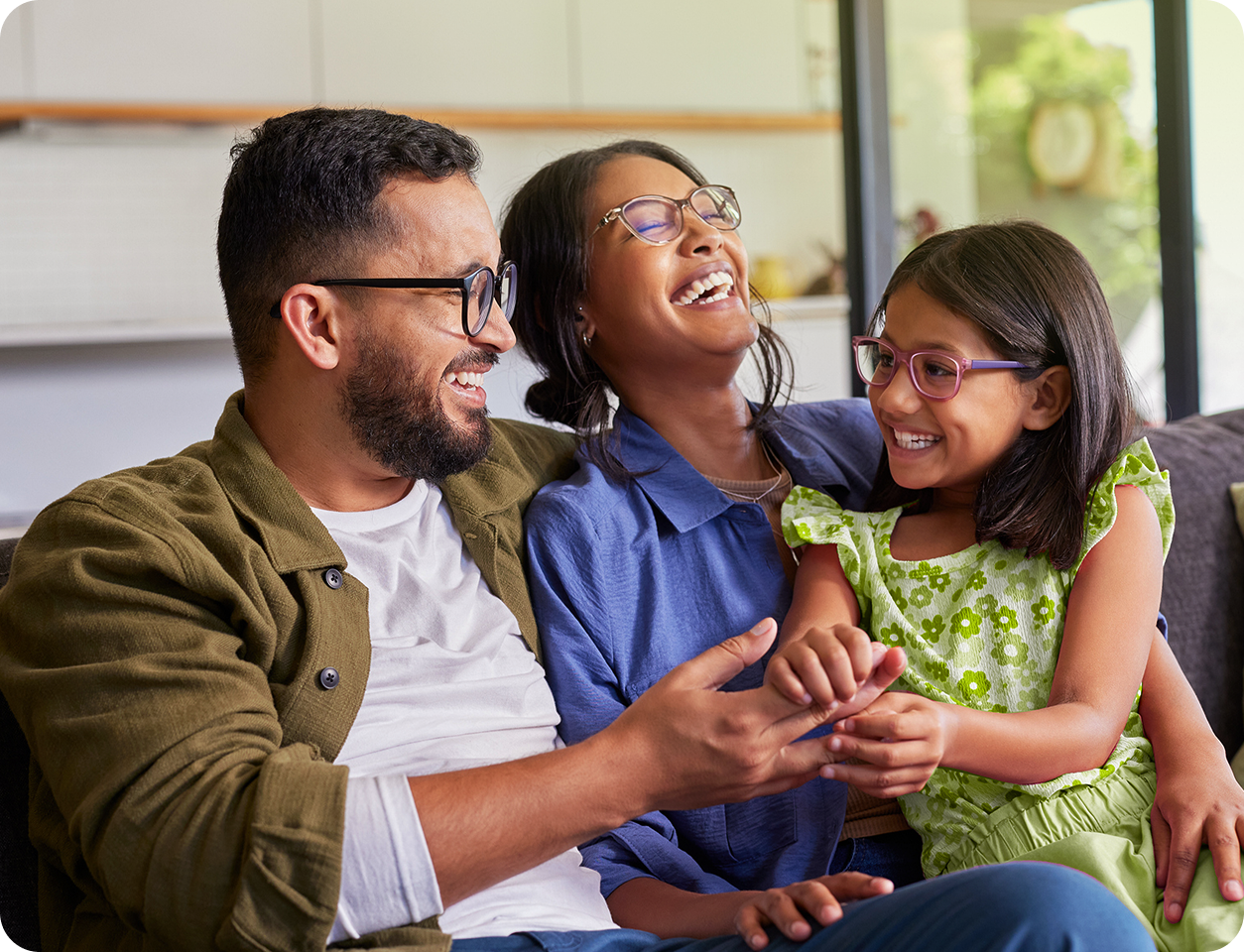 Família rindo junta dentro de casa. Pai e filha abraçam uma mulher no sofá, todos de óculos.