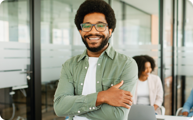 Homem negro sorridente, de óculos e braços cruzados, em um escritório moderno.